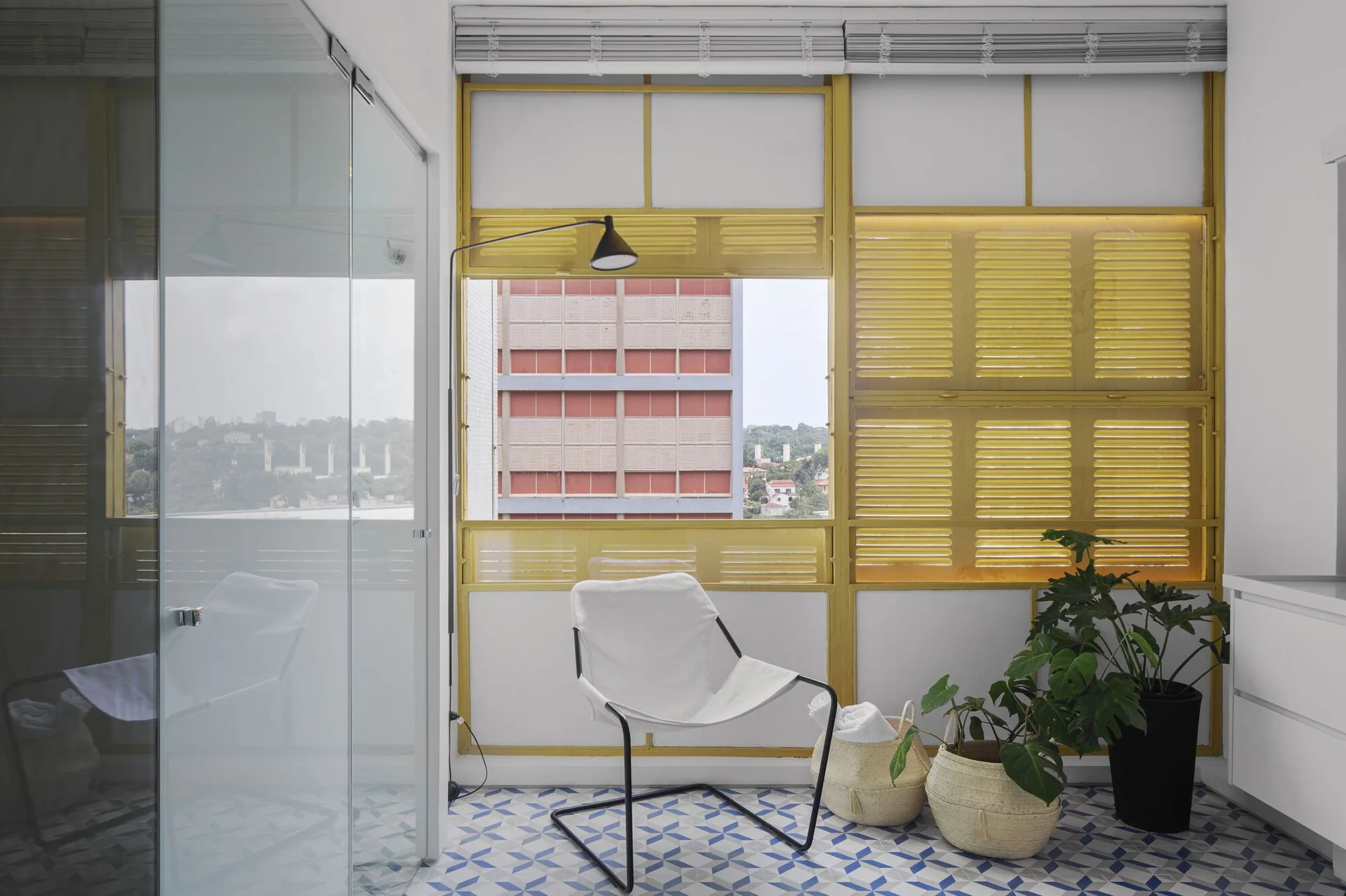 A modern sunroom features a white armchair, a blue and white geometric tile floor, and large yellow louvered window shutters.