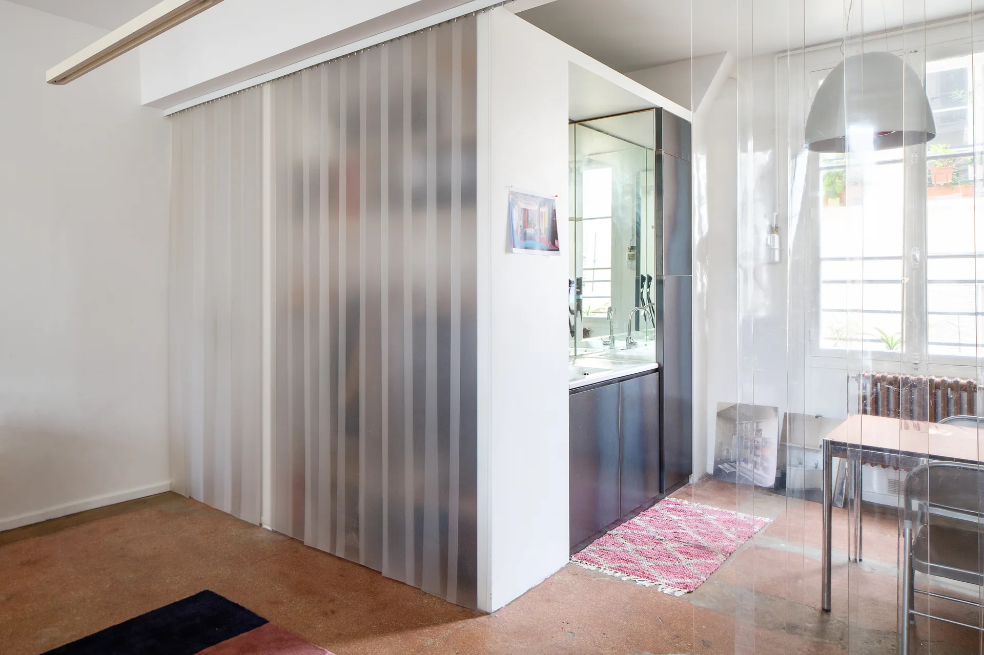 A bright industrial studio workspace features a translucent plastic strip curtain partition next to a vanity alcove and a minimalist metal desk.