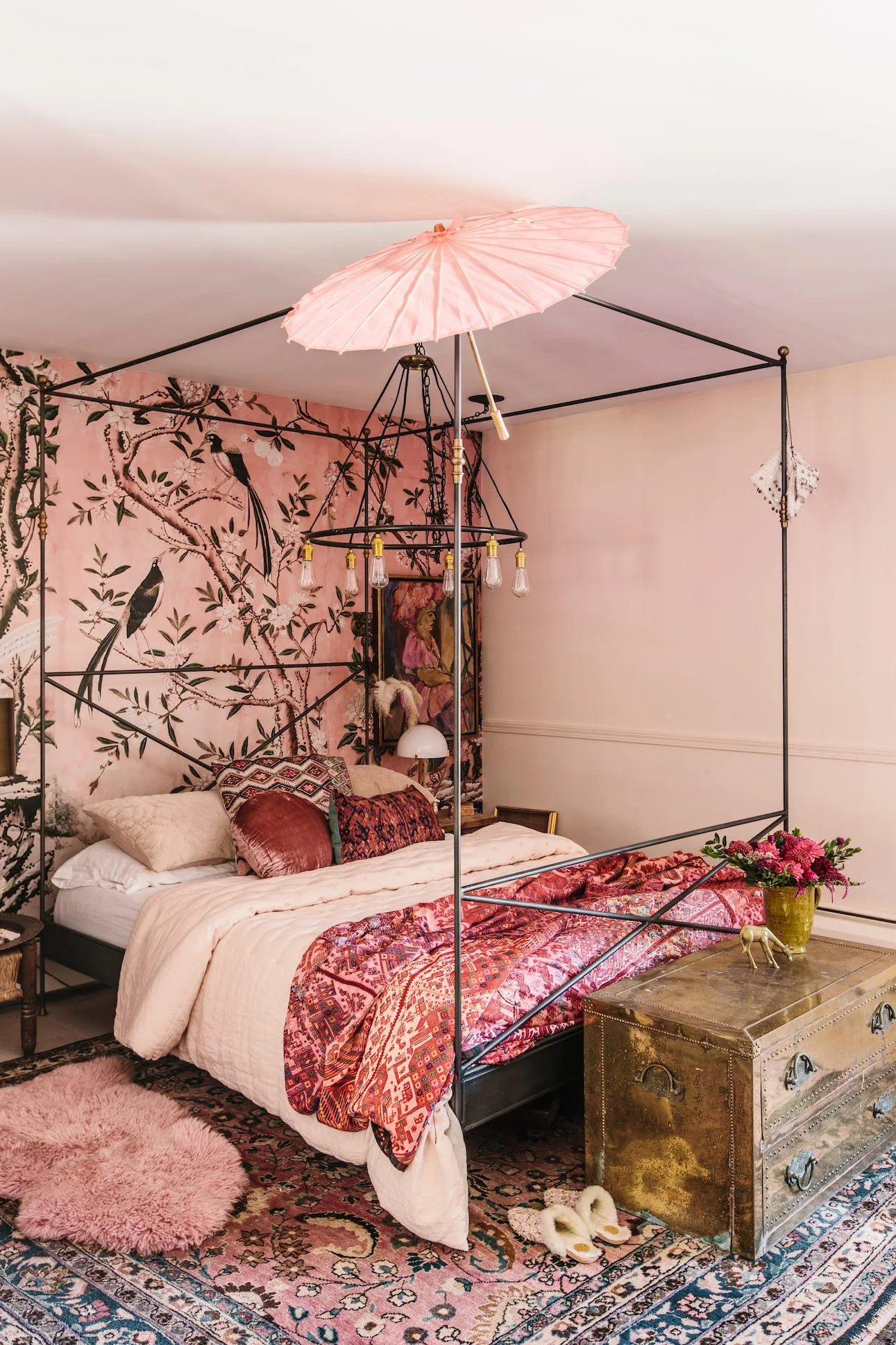 An eclectic pink bedroom features a black metal four-poster bed, botanical bird-print wallpaper, and a distressed brass trunk at the foot of the bed.