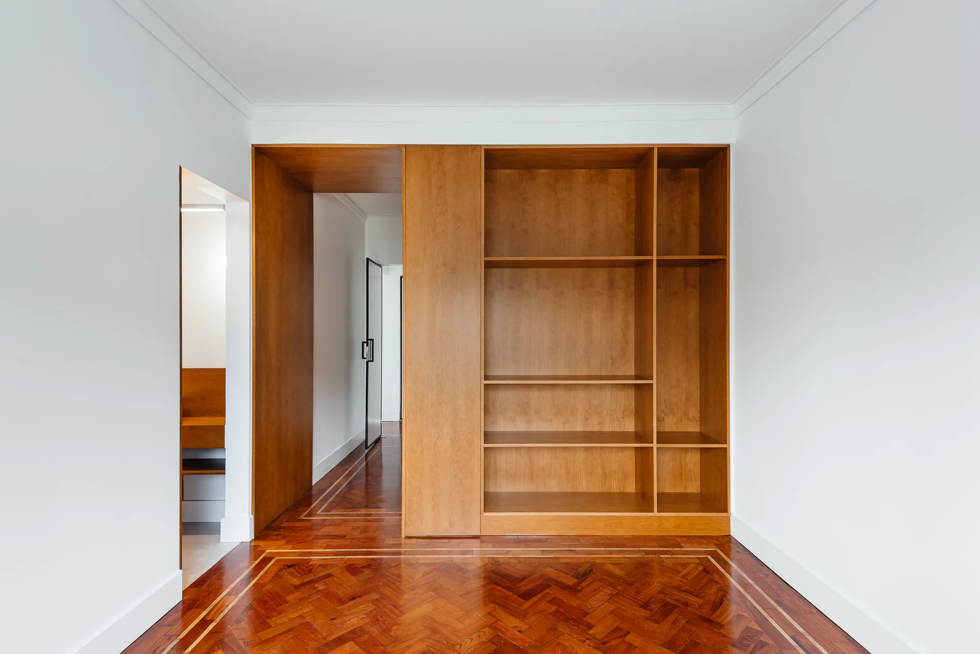A minimalist hallway features a tall built-in wooden shelving unit, a matching wood-paneled doorway, and a warm herringbone parquet floor with inlaid borders.