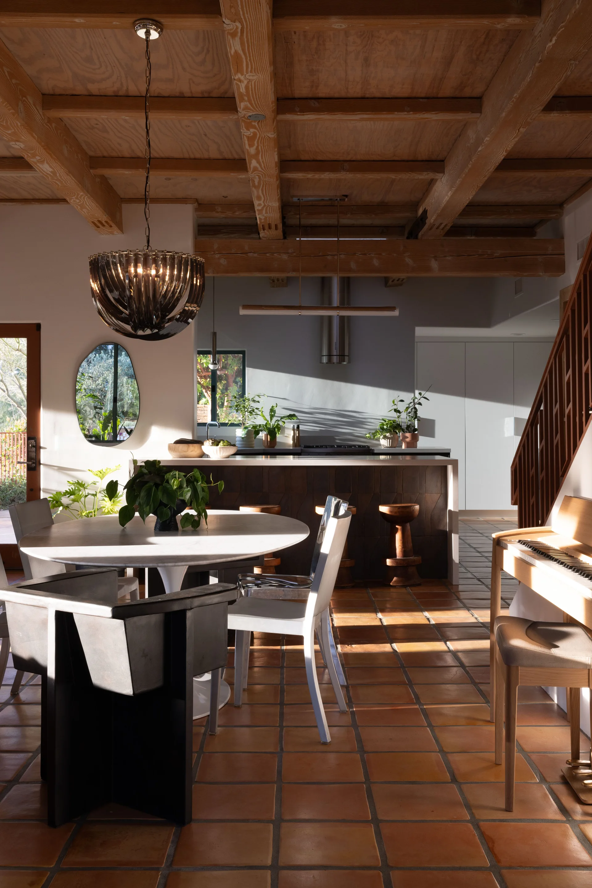 An open dining area features a white circular table with eclectic chairs, terracotta tile flooring, and a rustic exposed wood beam ceiling.