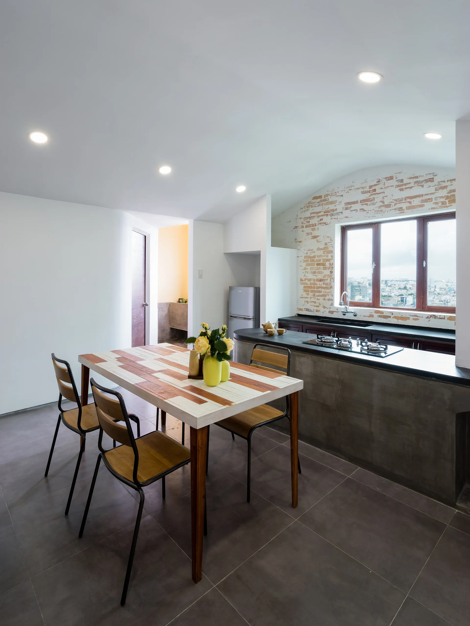 A modern industrial kitchen and dining area features a patterned wood table with chairs, a raw concrete kitchen island, and an exposed brick accent wall.