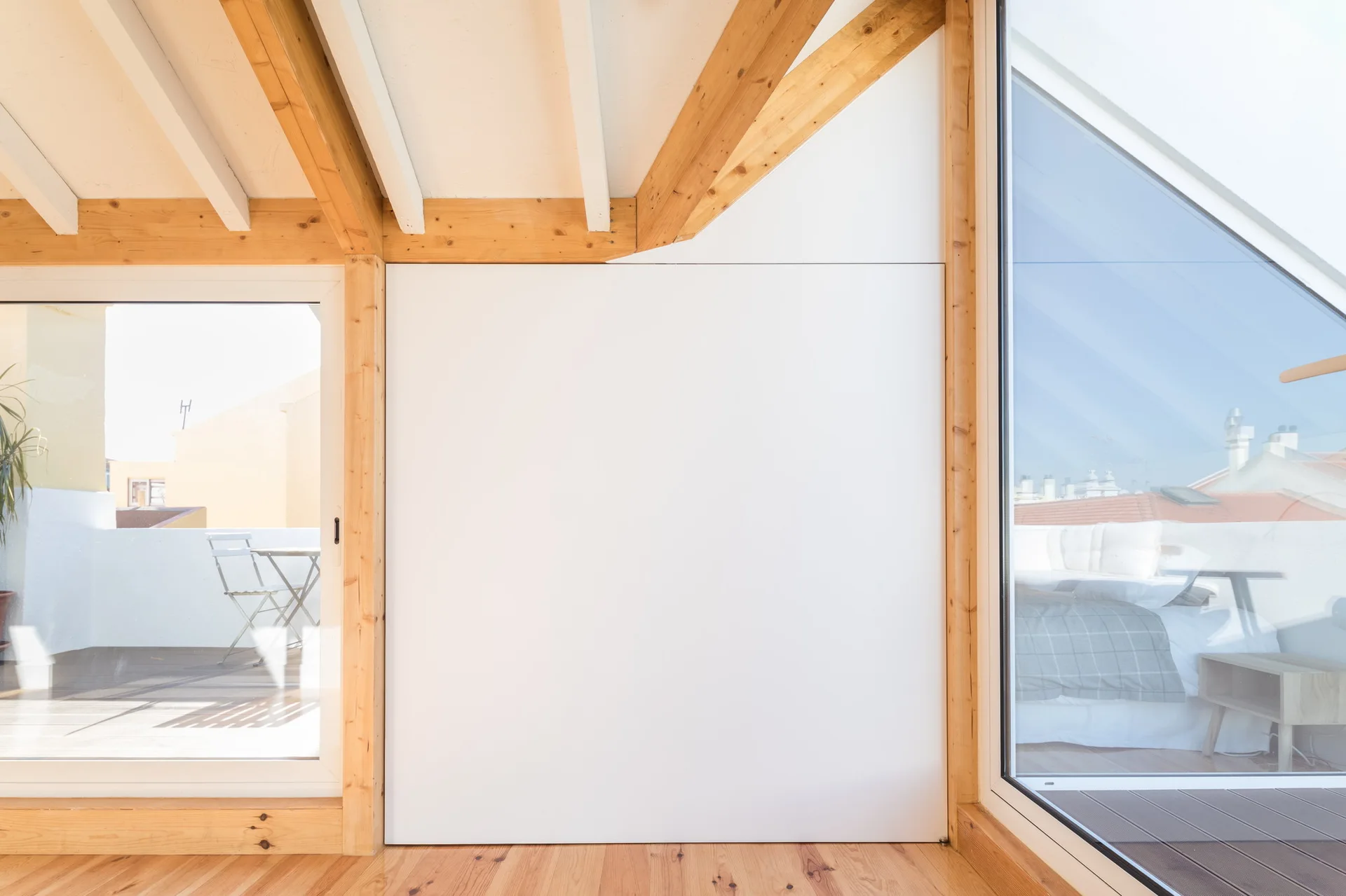 A minimalist attic interior with a central white wall panel flanked by large glass terrace doors under exposed natural timber ceiling beams.