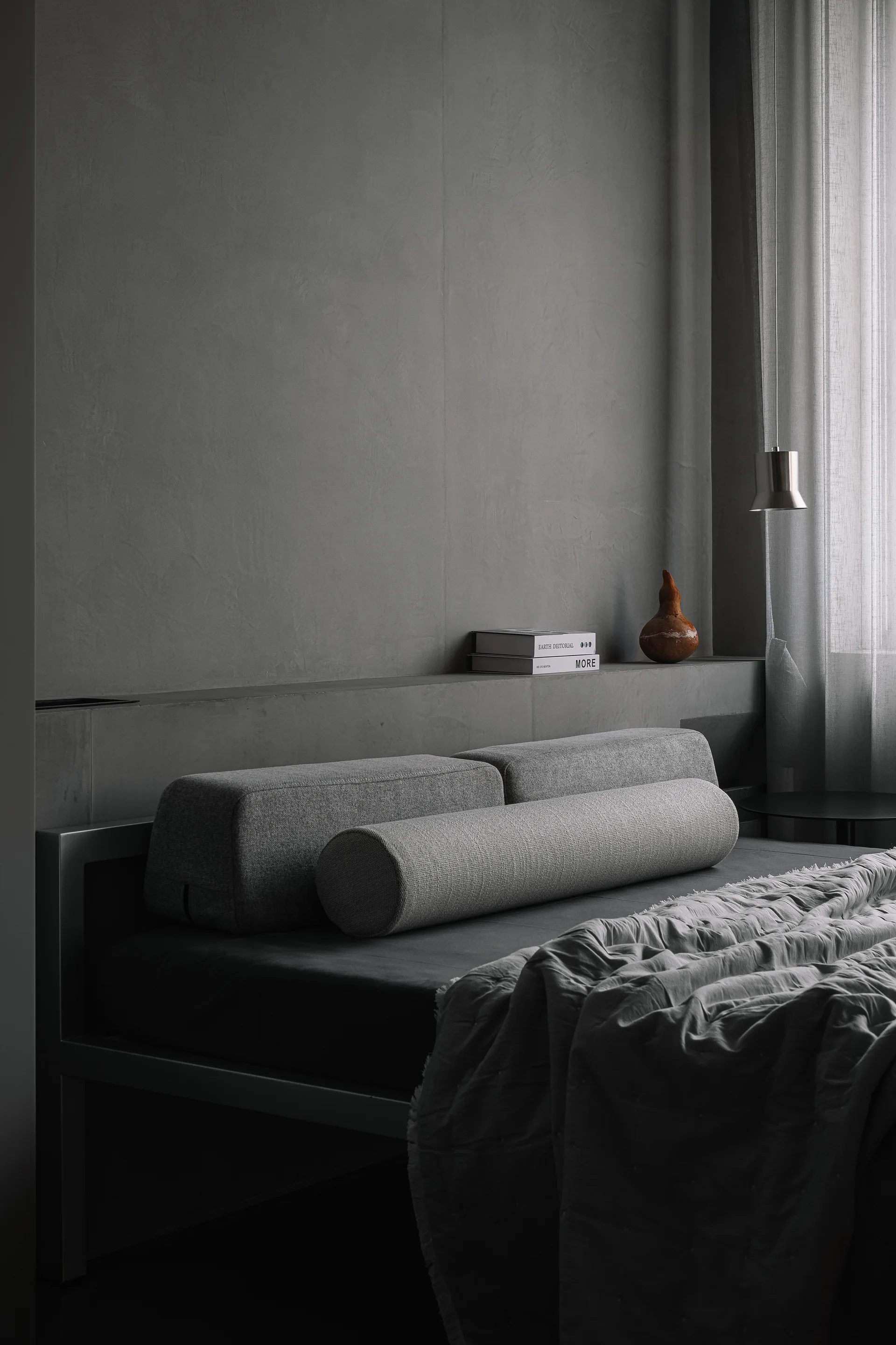 A minimalist grey bedroom features a low-profile bed with bolster pillows against a concrete ledge headboard and textured grey wall.
