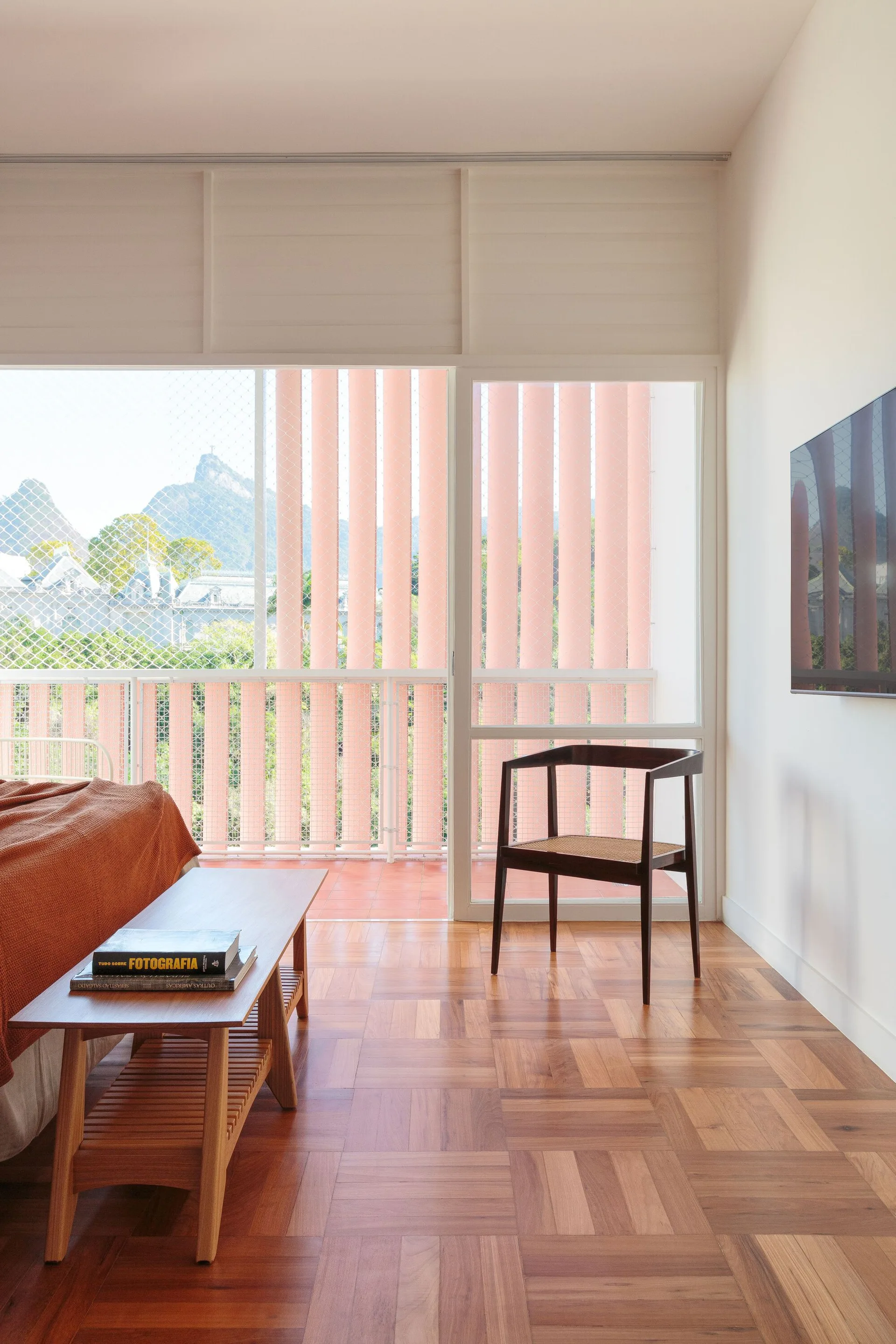 A minimalist bedroom features warm parquet wood flooring, a wooden bed bench, a dark wood chair, and large glass doors opening to a balcony.
