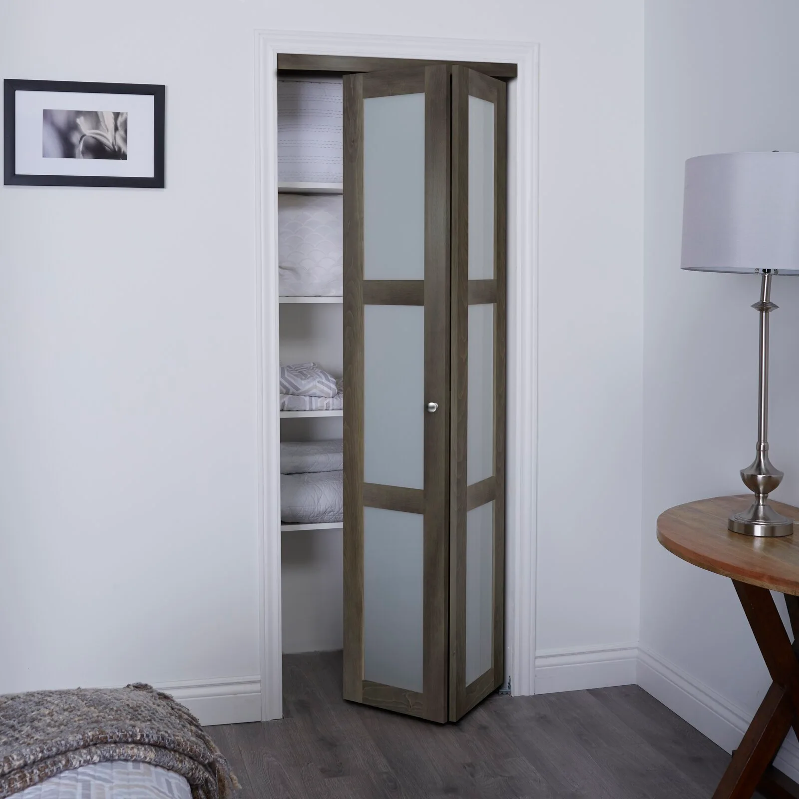 A grey wooden bifold door with frosted glass panels is partially open, revealing white closet shelves with folded linens next to a bedside lamp.