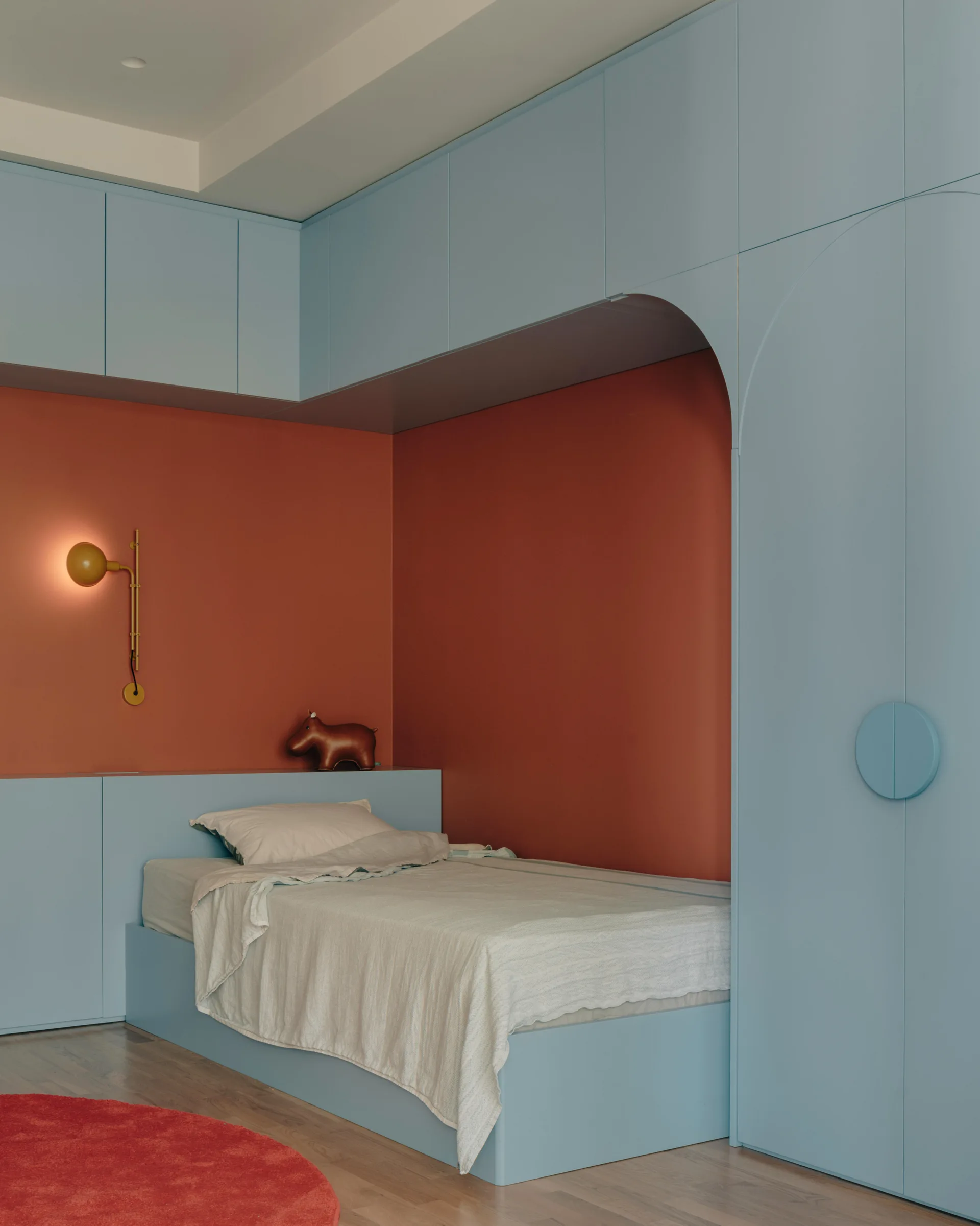 A light blue custom built-in wardrobe and cabinetry frame a single bed set against a matte burnt orange wall with a yellow globe wall sconce.