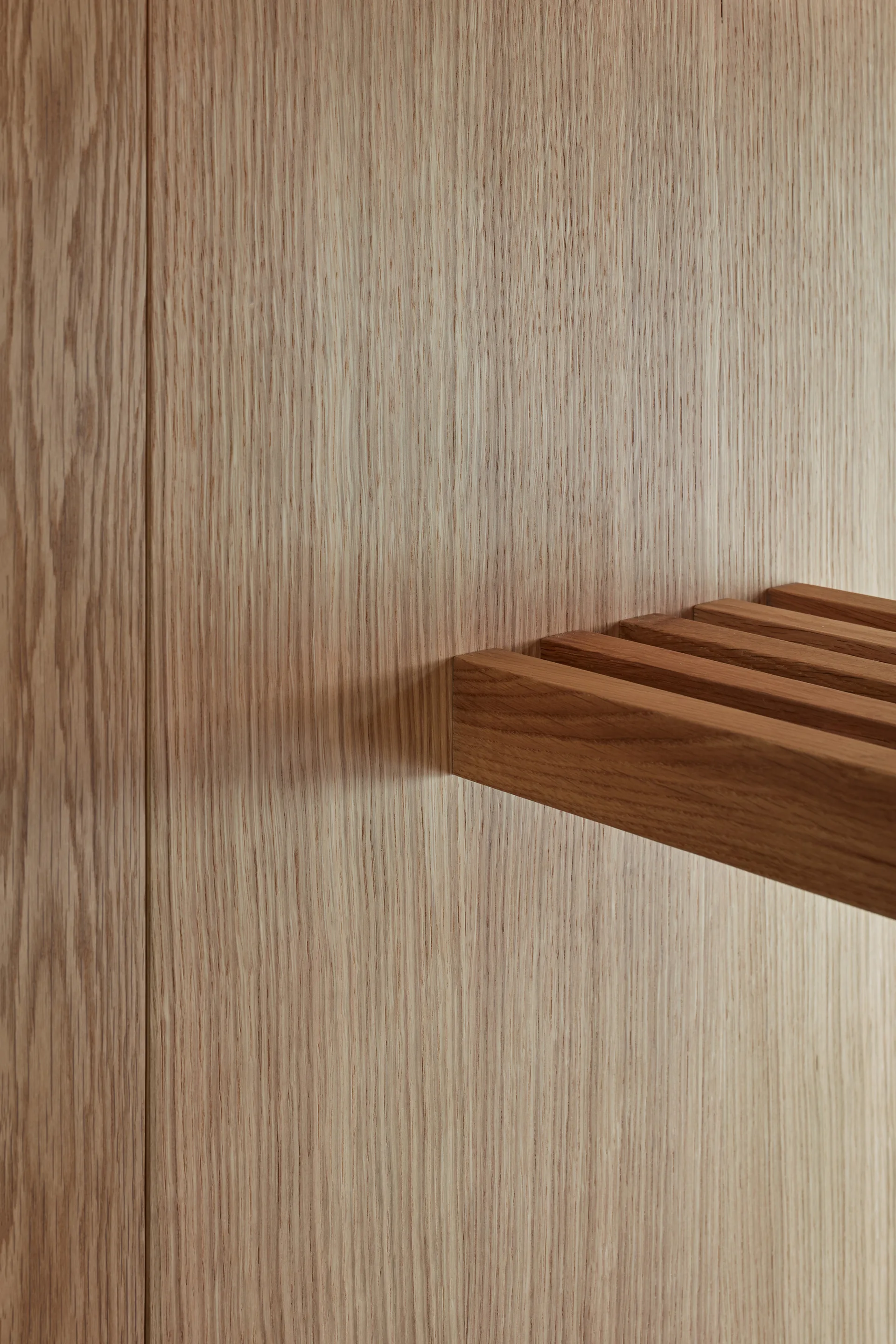 A close-up of a horizontal slatted wooden shelf cantilevered from a vertical grain light wood wall panel.