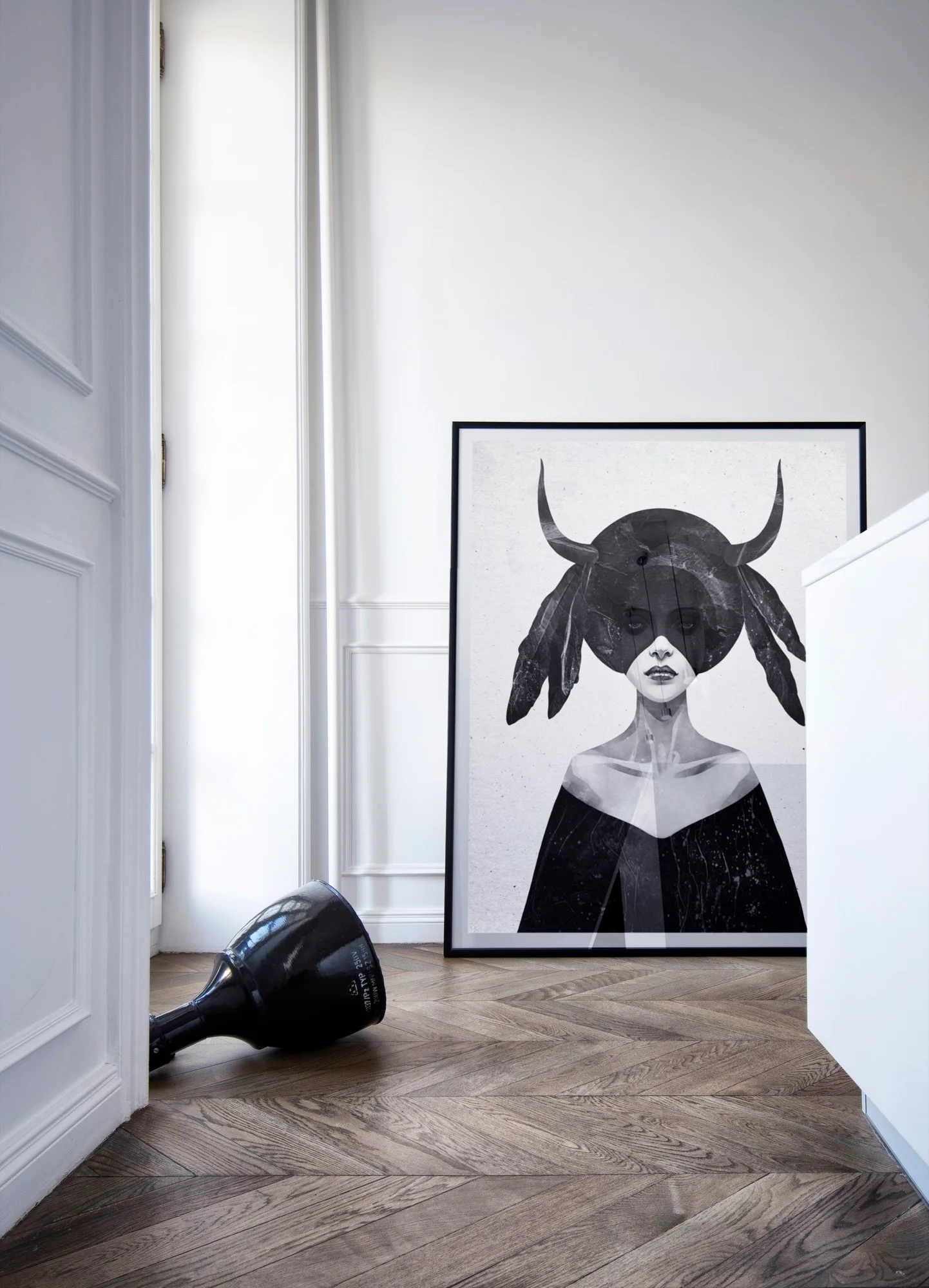 A large black and white framed art print leans against a white paneled wall above a herringbone patterned wood floor.
