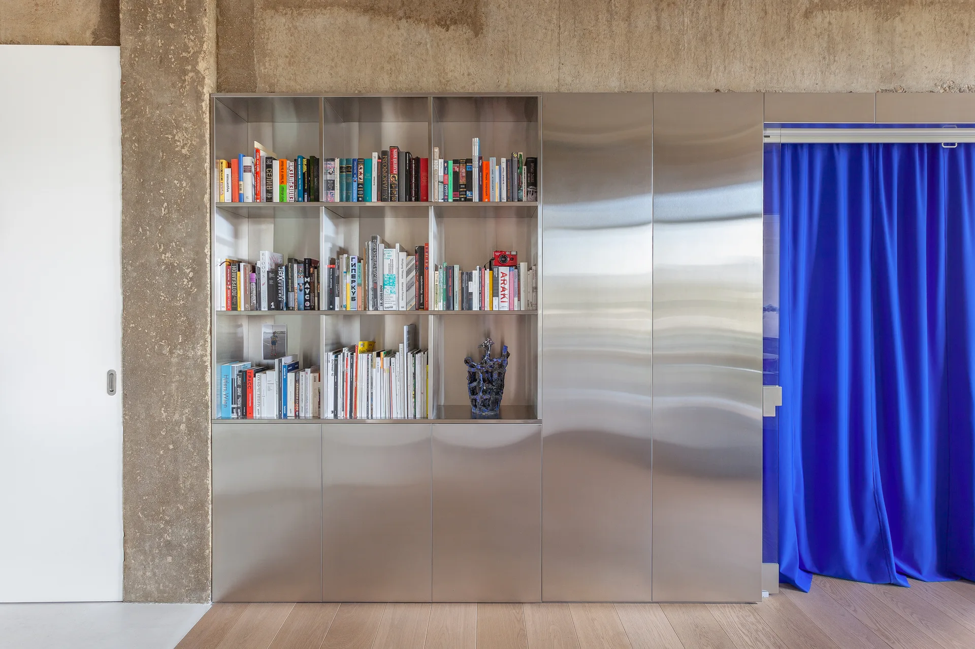 A brushed stainless steel built-in bookshelf and cabinet unit stands against a textured concrete wall next to a vibrant blue fabric curtain.