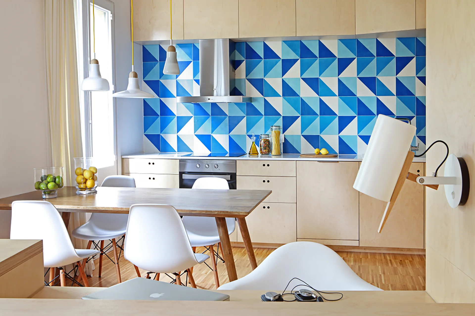 A bright kitchen features light plywood cabinets, a blue and white geometric tile backsplash, and a wooden dining table with white plastic chairs.