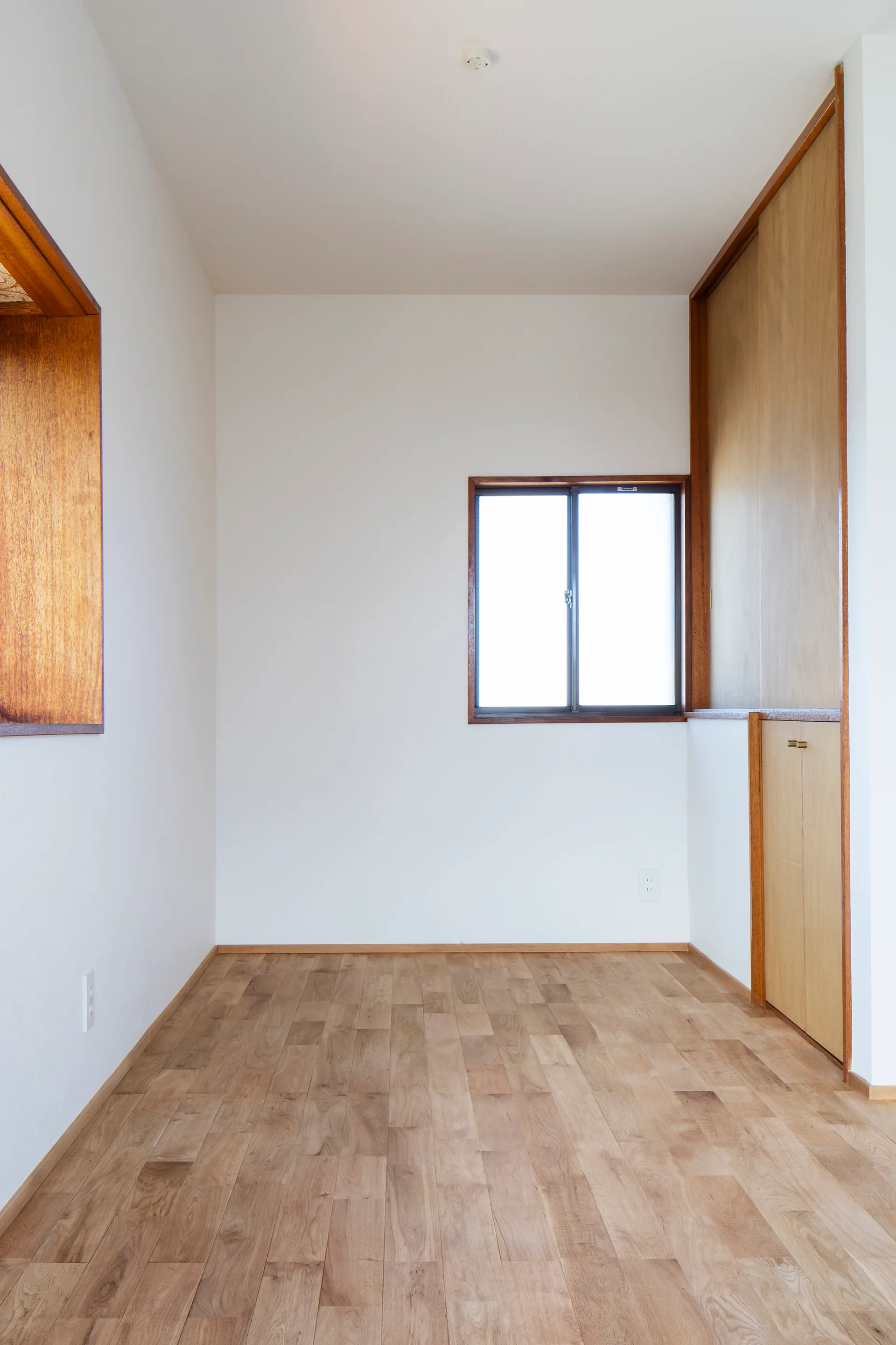 An empty minimalist room featuring white walls, light wood plank floors, a small window with wood trim, and a built-in wooden closet.