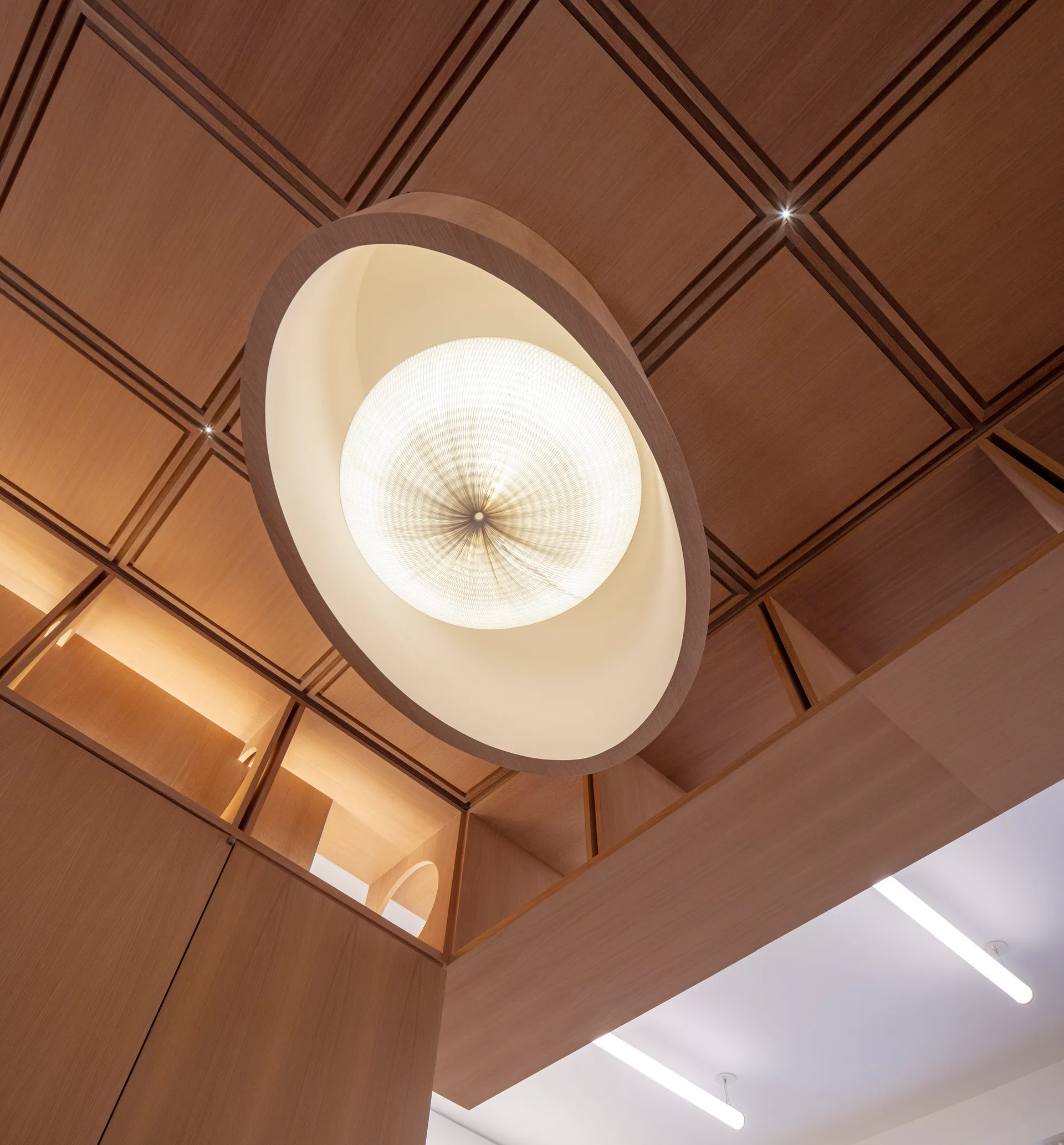 A low-angle view of a large circular pendant light set in a wooden frame below a wood-paneled coffered ceiling with built-in cabinetry.