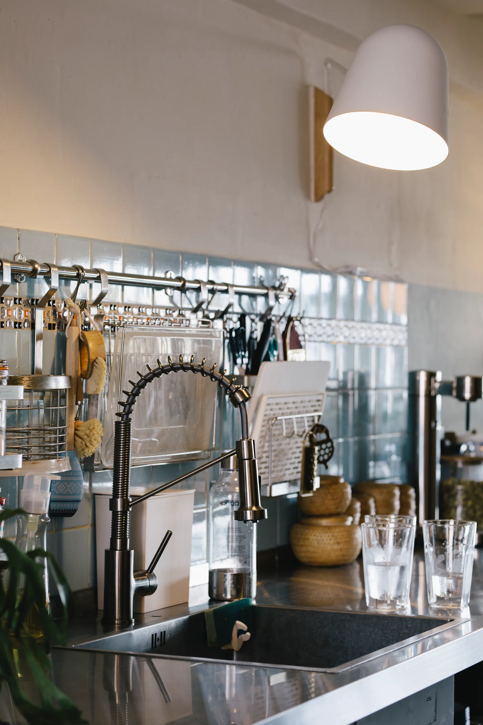 A stainless steel kitchen sink features a commercial spring faucet set against a glossy blue tiled backsplash with a wall-mounted utensil hanging rail.