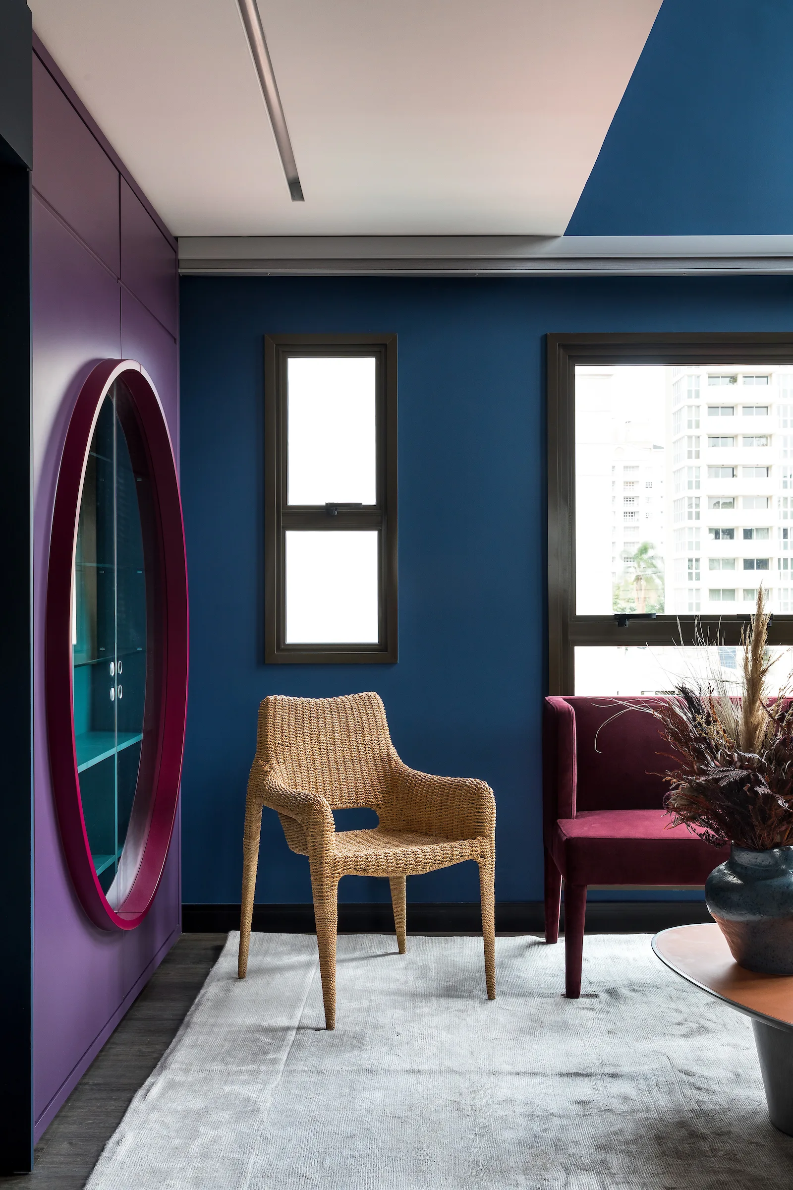 A modern living room features a blue wall, a purple built-in cabinet with a circular display window, and a woven rattan armchair on a grey rug.