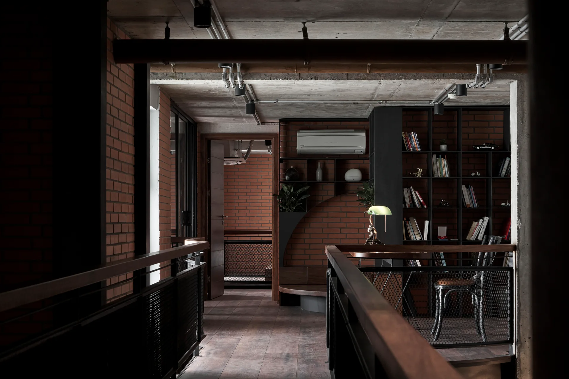 An industrial loft landing features exposed red brick walls, a raw concrete ceiling with metal pipes, and a dark metal shelving unit with a green banker's lamp.