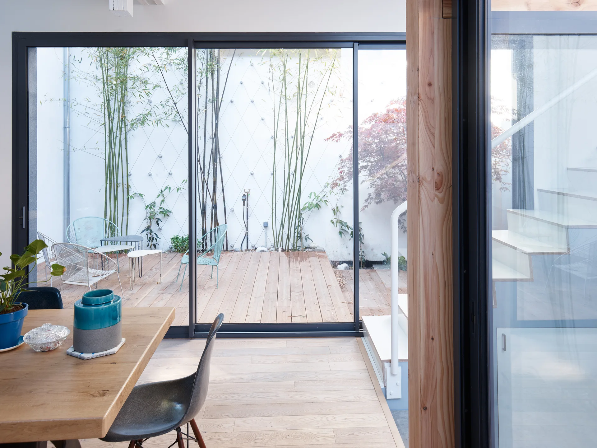 A wooden dining table looks out through large sliding glass doors to a wooden patio deck lined with tall bamboo and metal chairs.