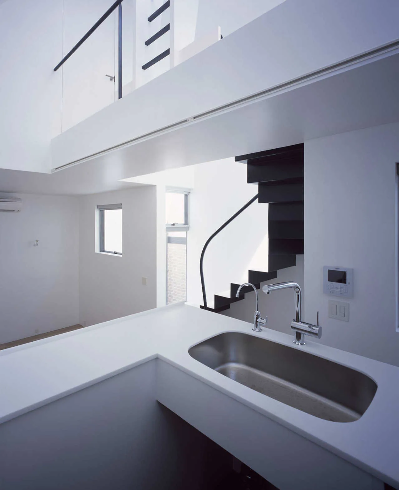 A minimalist white kitchen counter with a stainless steel sink sits in front of a modern black staircase in a bright split-level home.