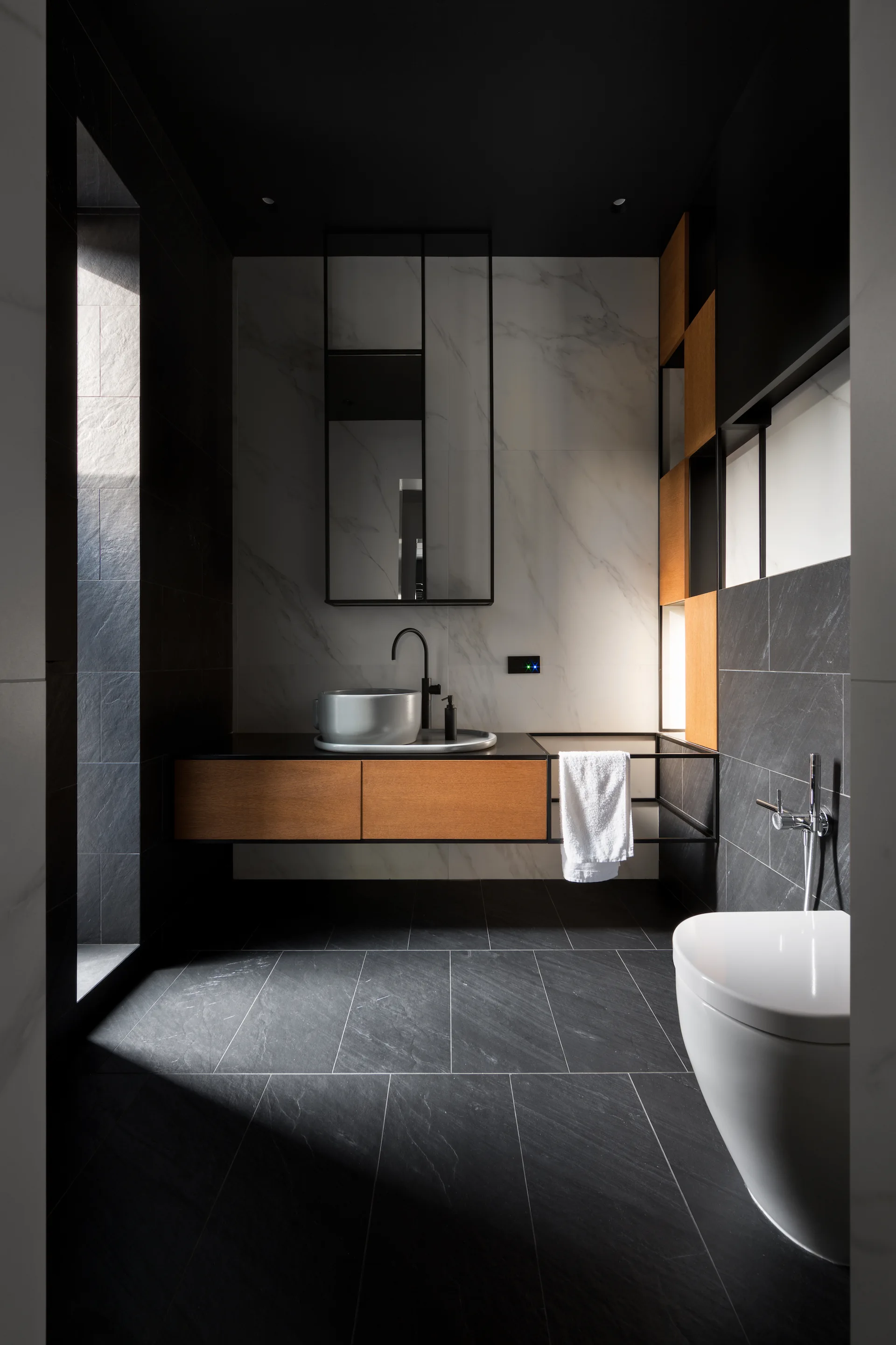 A modern minimalist bathroom featuring a floating wooden vanity, a light gray vessel sink, and dark slate floor tiles.