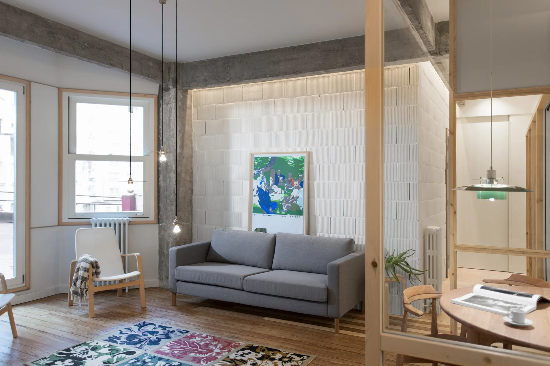 A grey sofa sits against a white block wall below concrete ceiling beams, accented by a colorful patterned rug on wood floors.