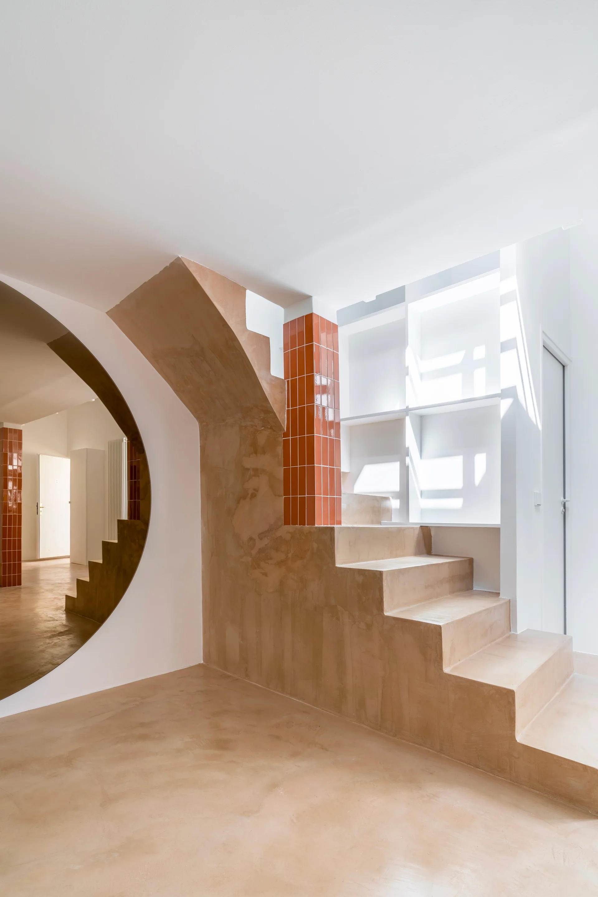 A minimalist plaster staircase in warm beige features a glossy terracotta tiled column and a circular cutout mirror.