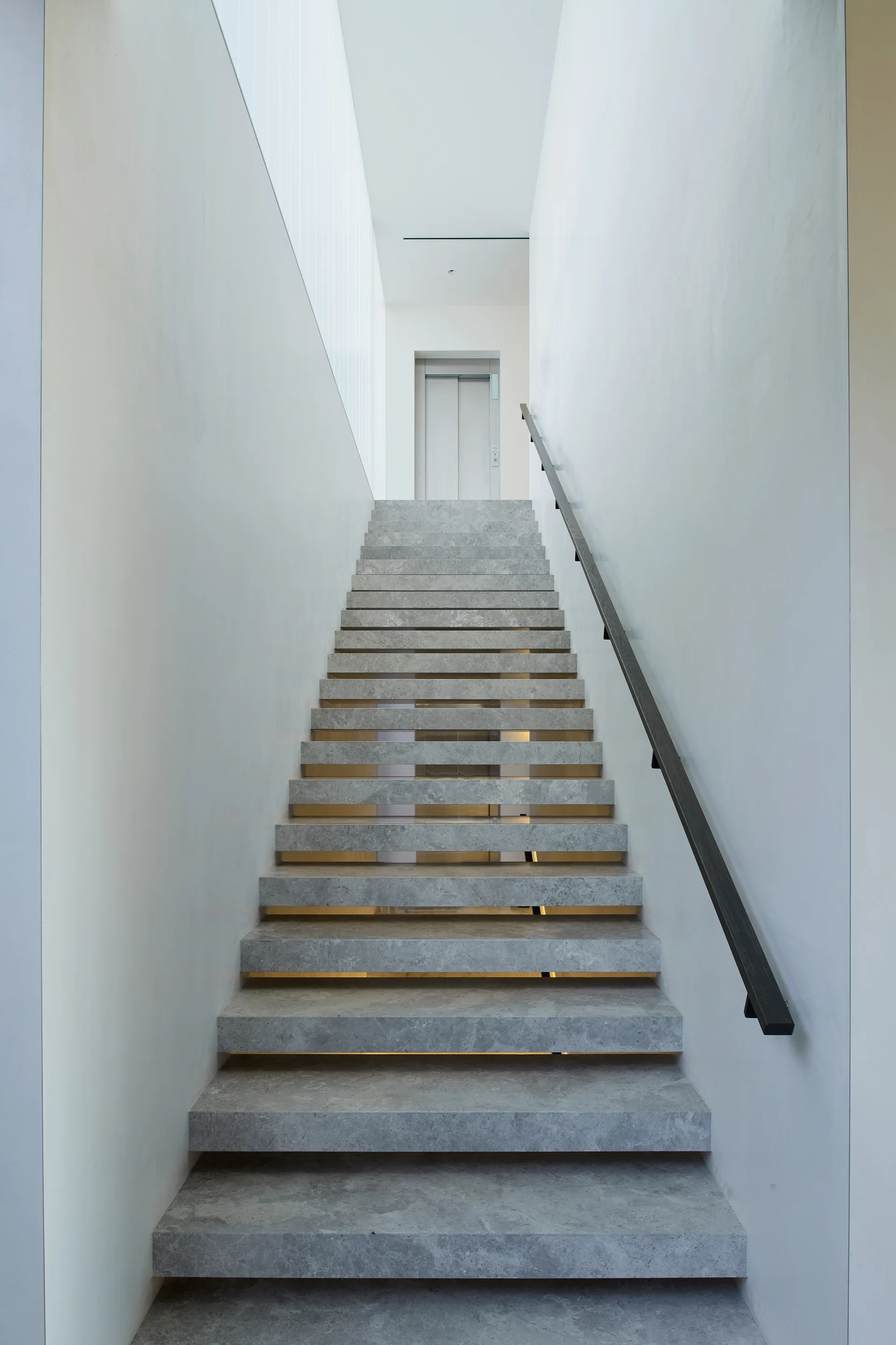 A contemporary staircase features grey stone steps with warm under-tread lighting and a black handrail set between smooth white walls.