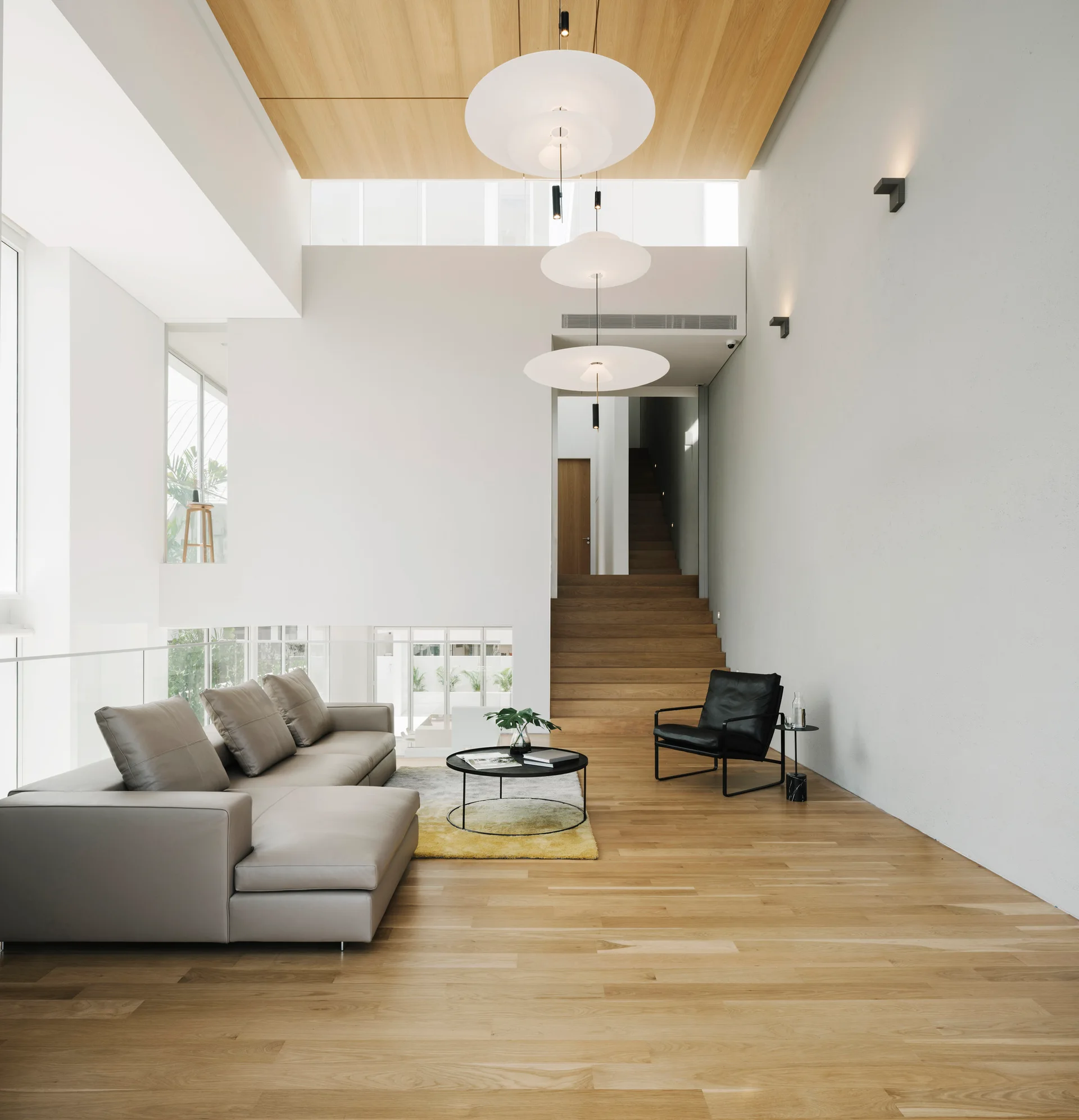A modern double-height living room featuring a grey sectional sofa, a black armchair, wood-plank flooring, and stacked white disk pendant lights.