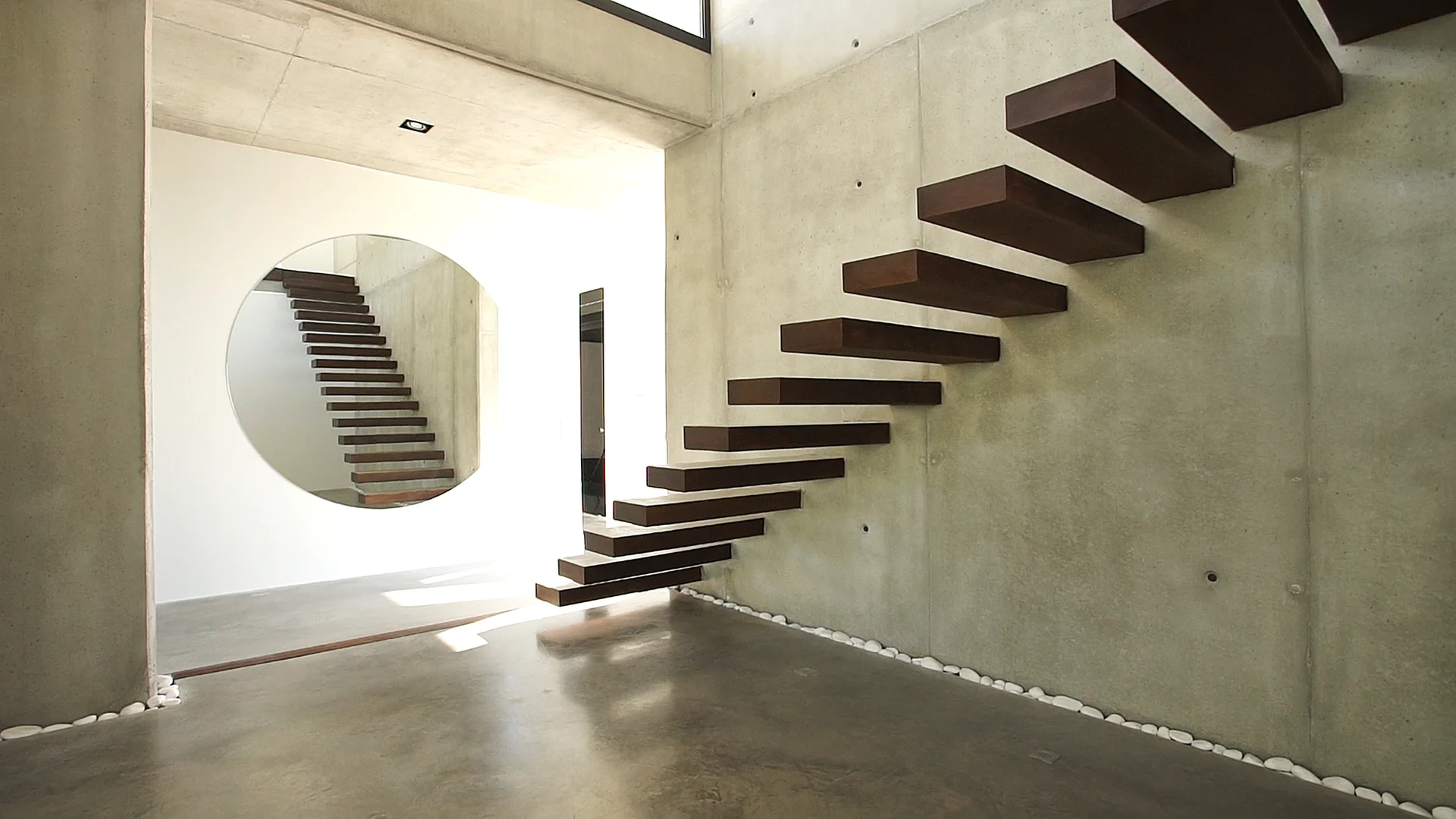 A cantilevered wooden staircase climbs a raw concrete wall adjacent to a large round mirror reflecting the steps above polished floors.