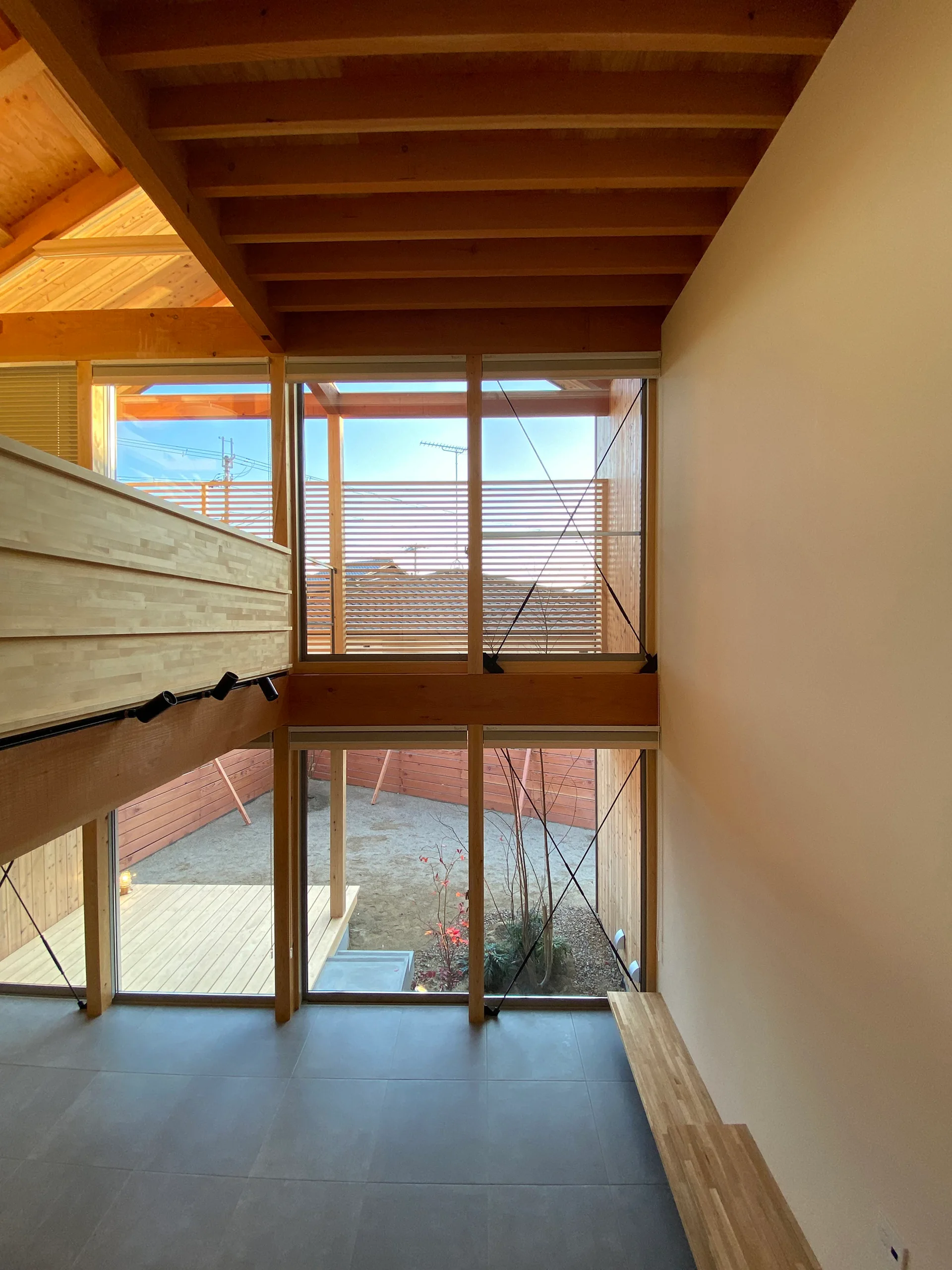 A double-height room features exposed wood ceiling beams, a large two-story window overlooking a courtyard, gray tiled flooring, and white walls.