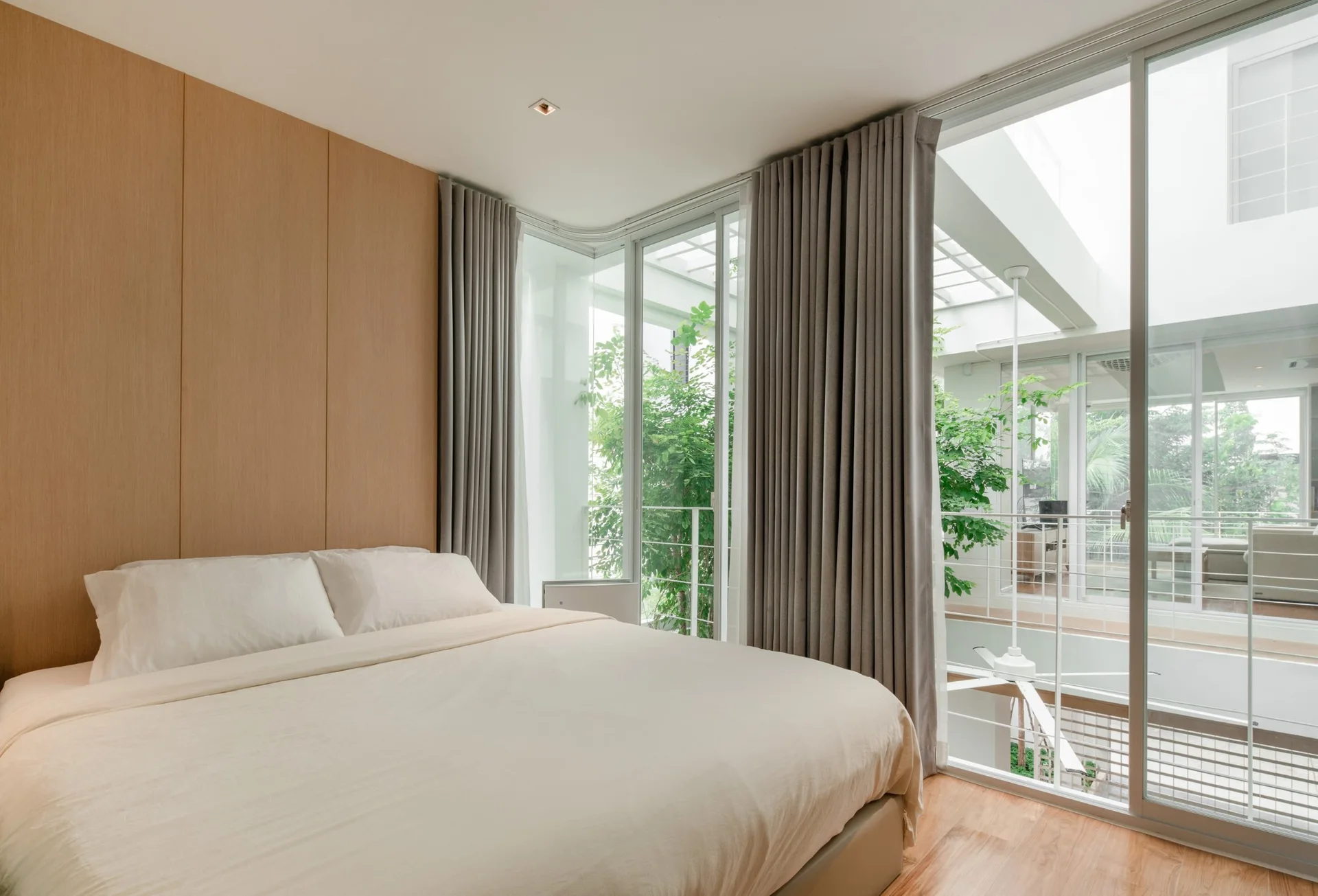 A minimalist bedroom features a bed against a wood-paneled wall, grey curtains, and large sliding glass doors opening to a courtyard.