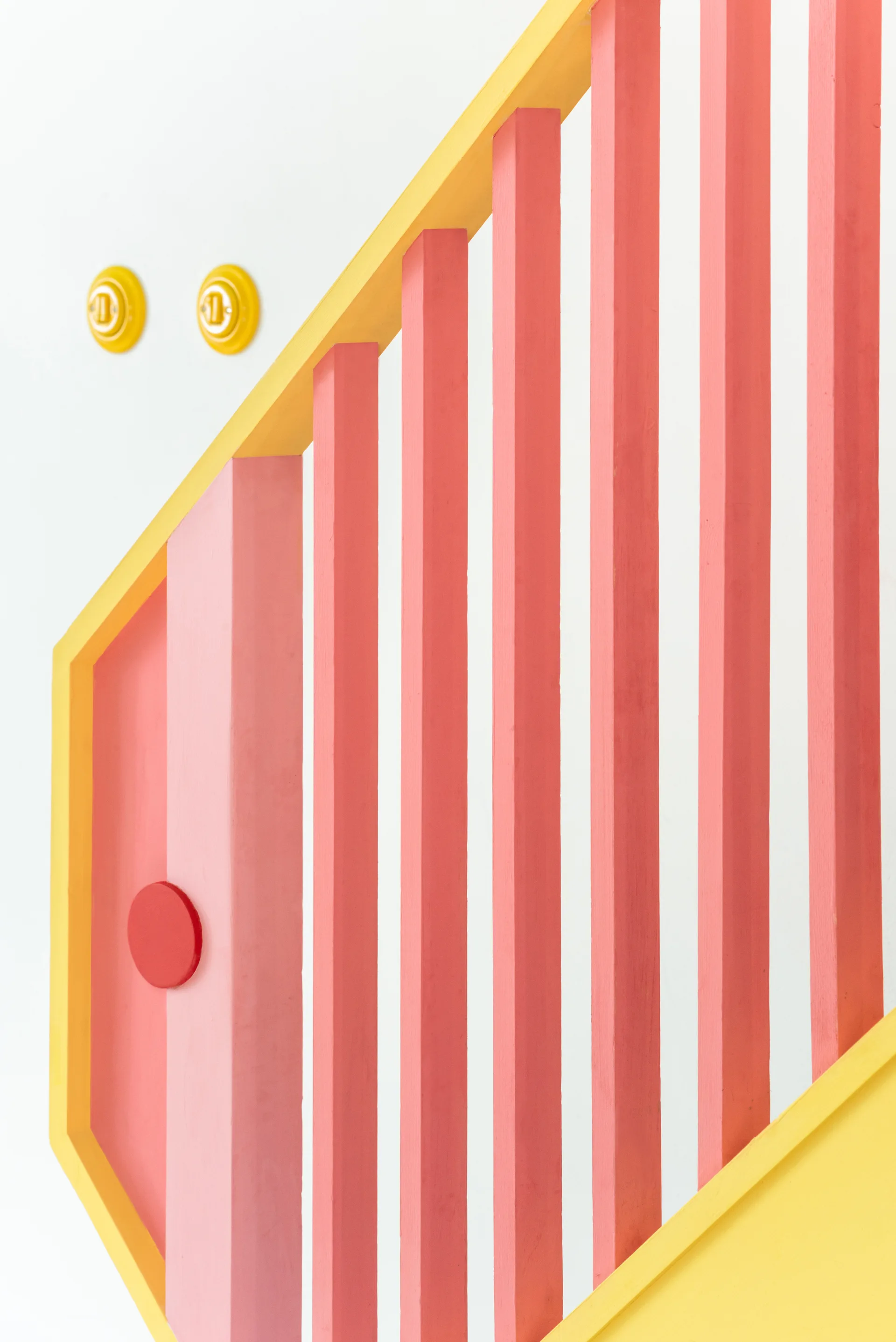 A close-up view of a modern wooden staircase railing painted with pink and red vertical balusters, a yellow handrail, and circular yellow wall switches.