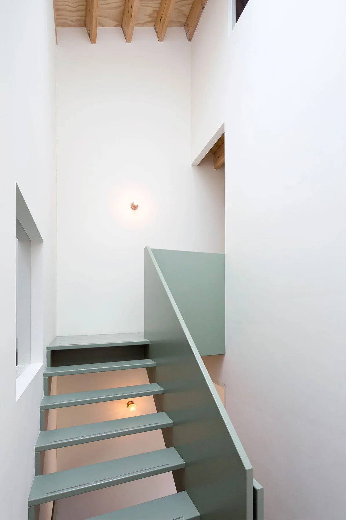 A minimalist sage green staircase with a solid balustrade rises against plain white walls under exposed wood ceiling beams.