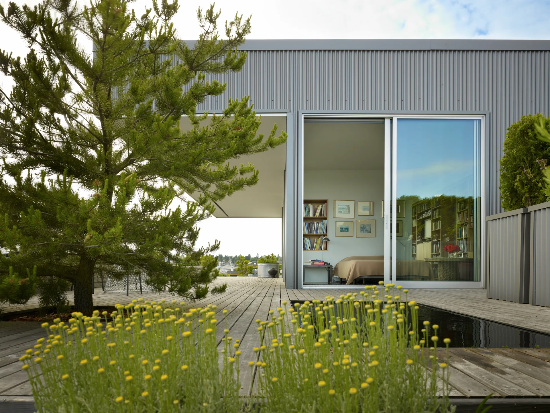 A modern outdoor terrace features weathered gray wooden decking, a sunken reflecting pool, corrugated metal siding, and a large sliding glass door.