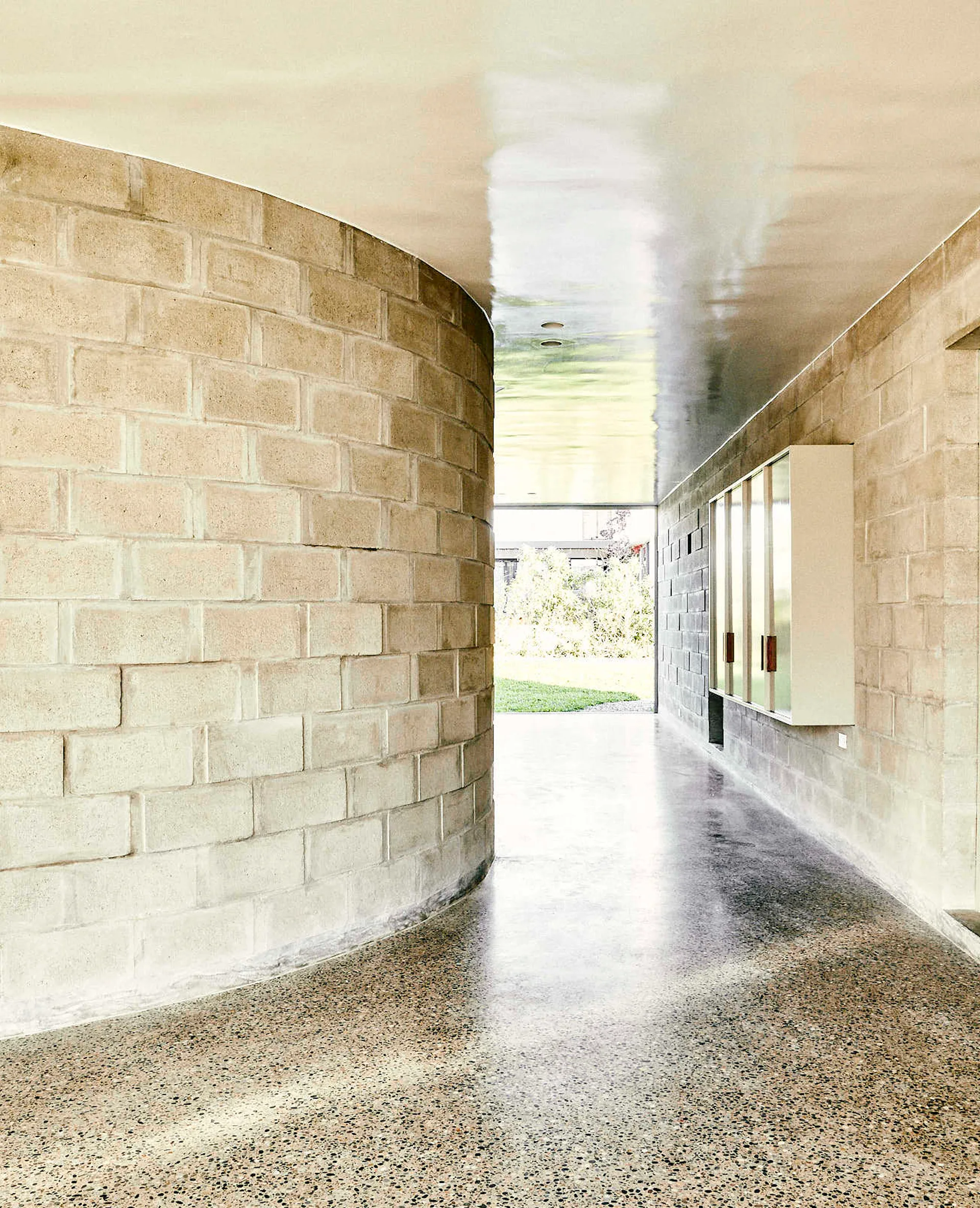 A minimalist hallway features a curved concrete block wall, a highly reflective glossy ceiling, and a polished speckled floor leading to a garden view.