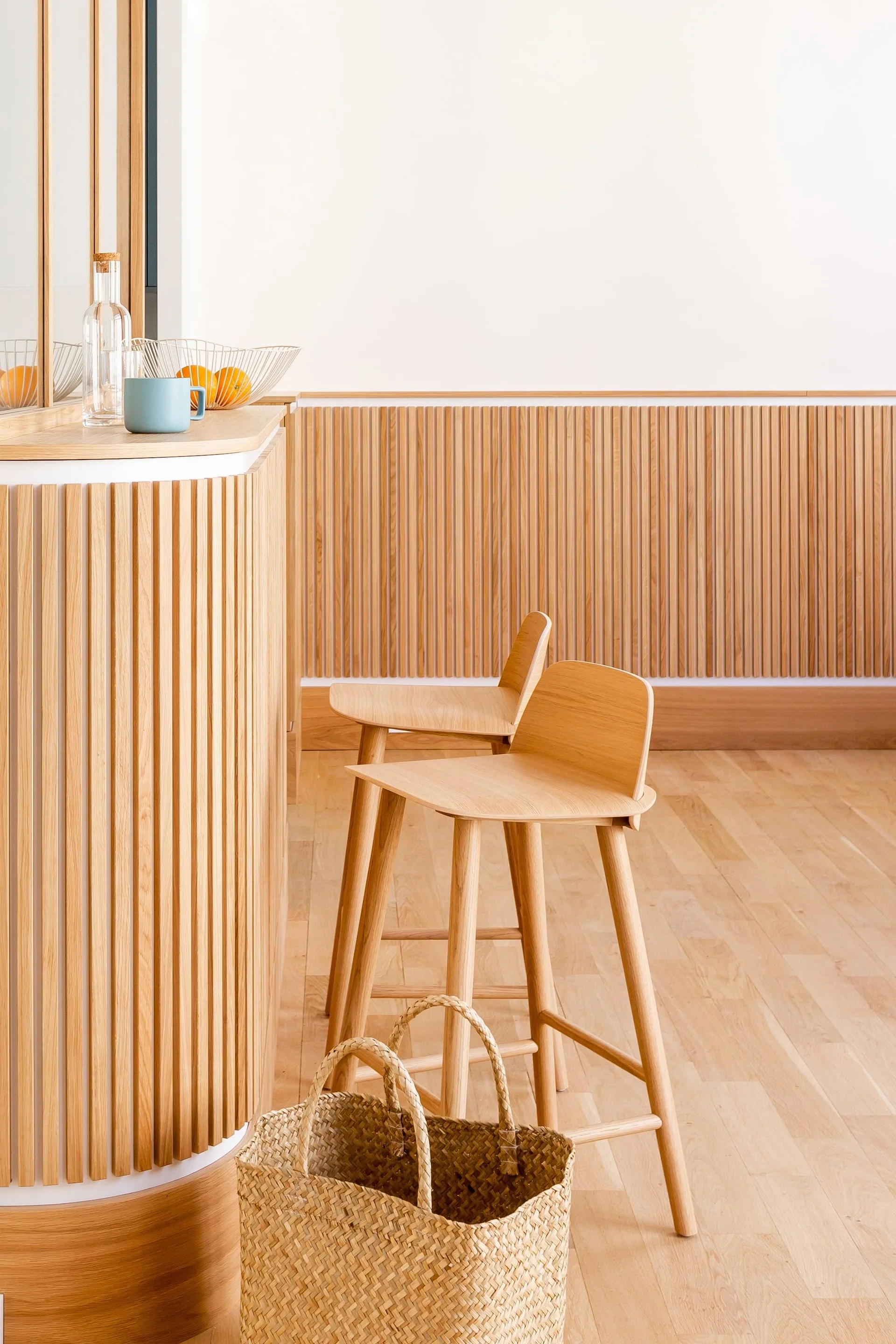 A curved light wood kitchen island with vertical slats features two matching wooden bar stools against a slatted wood paneled wall.