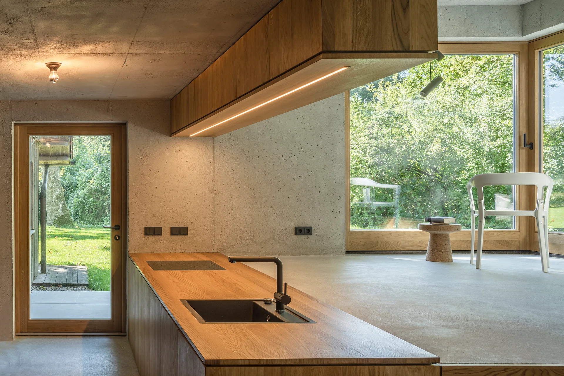 A minimalist kitchen features a wood-clad island and matching suspended overhead cabinets against textured raw concrete walls and large windows.