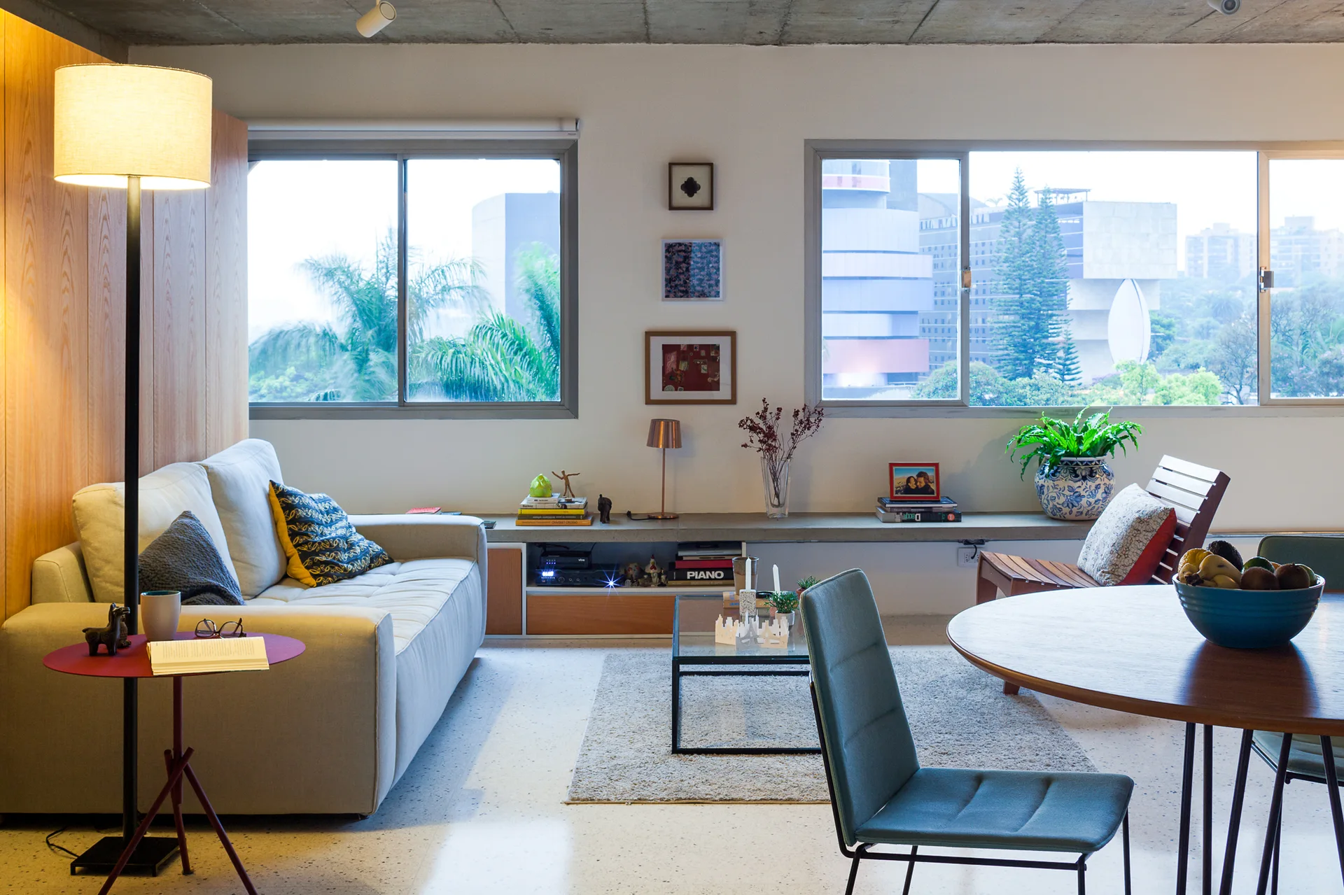 A modern living room features an exposed concrete ceiling, a light gray sofa, two large windows, and a low-slung built-in concrete shelf.