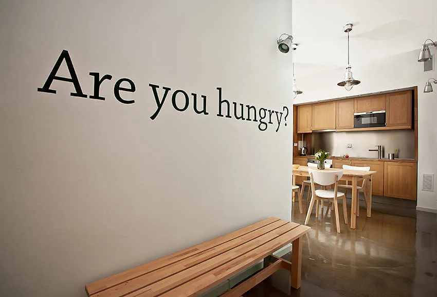 A wooden bench sits near a white wall featuring a black text decal that leads into a modern kitchen with natural wood cabinets.