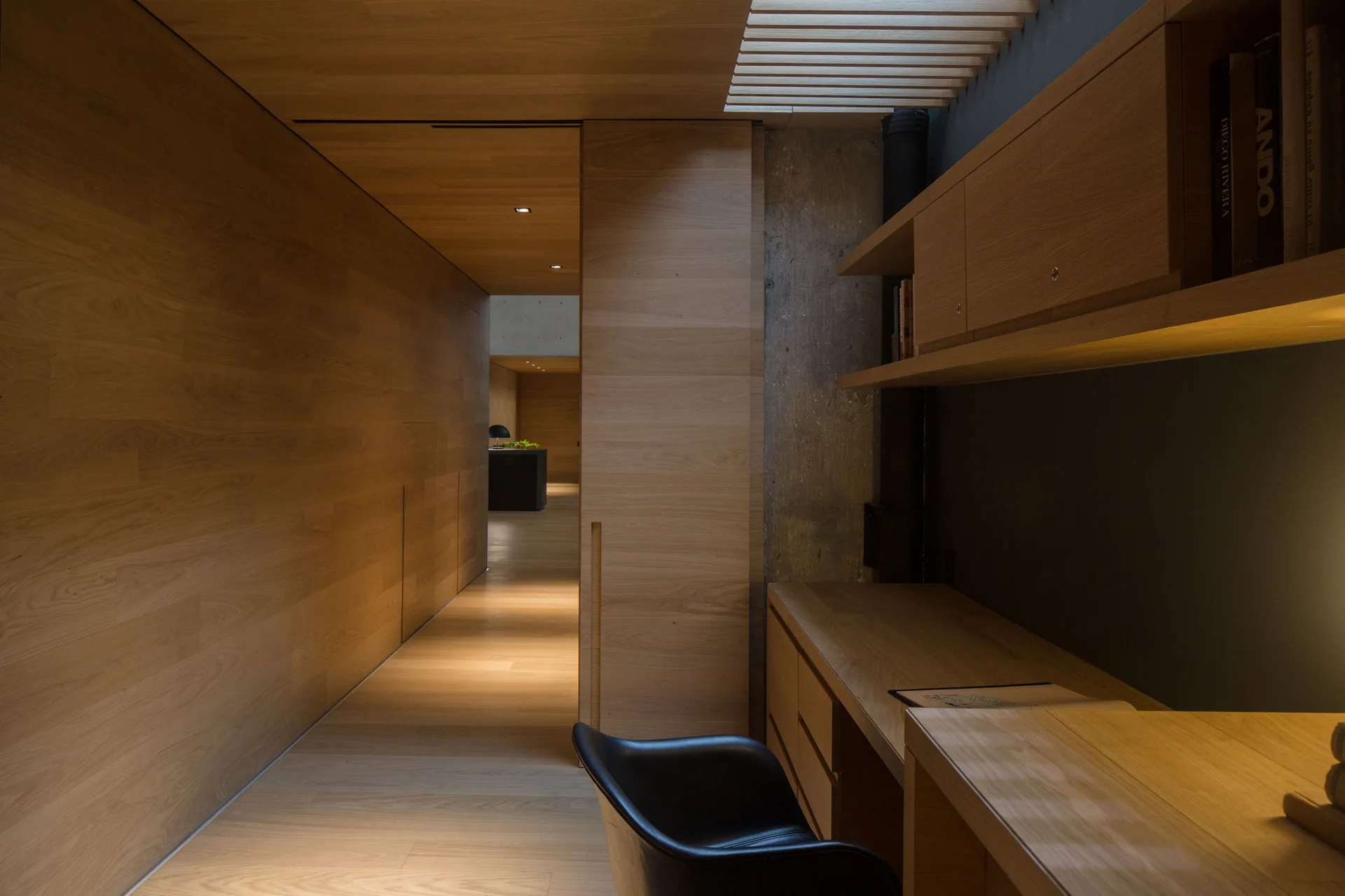 A wood-paneled home office nook featuring a built-in wooden desk, a black leather chair, an exposed concrete accent wall, and an overhead slatted skylight.