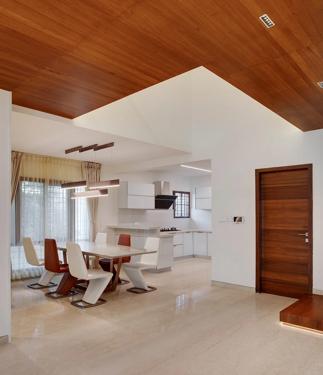 A modern dining area features a glossy stone table with white and rust chairs beneath a rich wood ceiling panel that flows into an adjacent kitchen.
