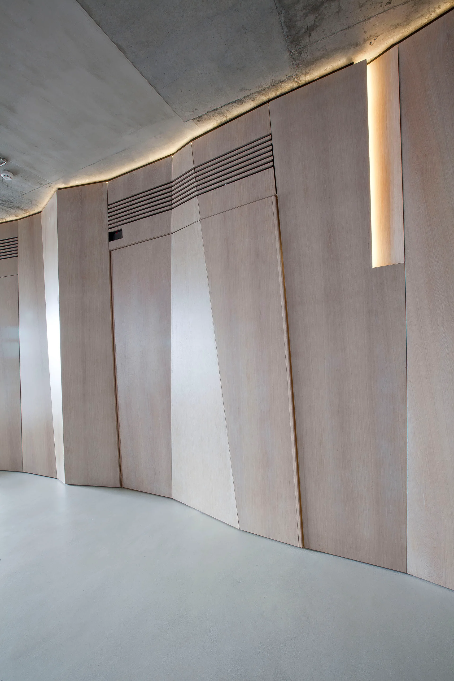 A curved wall clad in light wood veneer panels stands beneath a raw concrete ceiling with integrated cove lighting and horizontal air vents.
