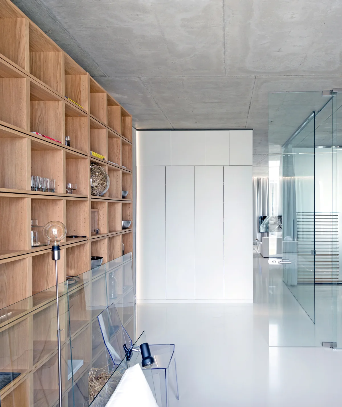 A modern hallway features a large oak grid bookshelf, white floor-to-ceiling cabinets, and a reflective white floor under a raw concrete ceiling.