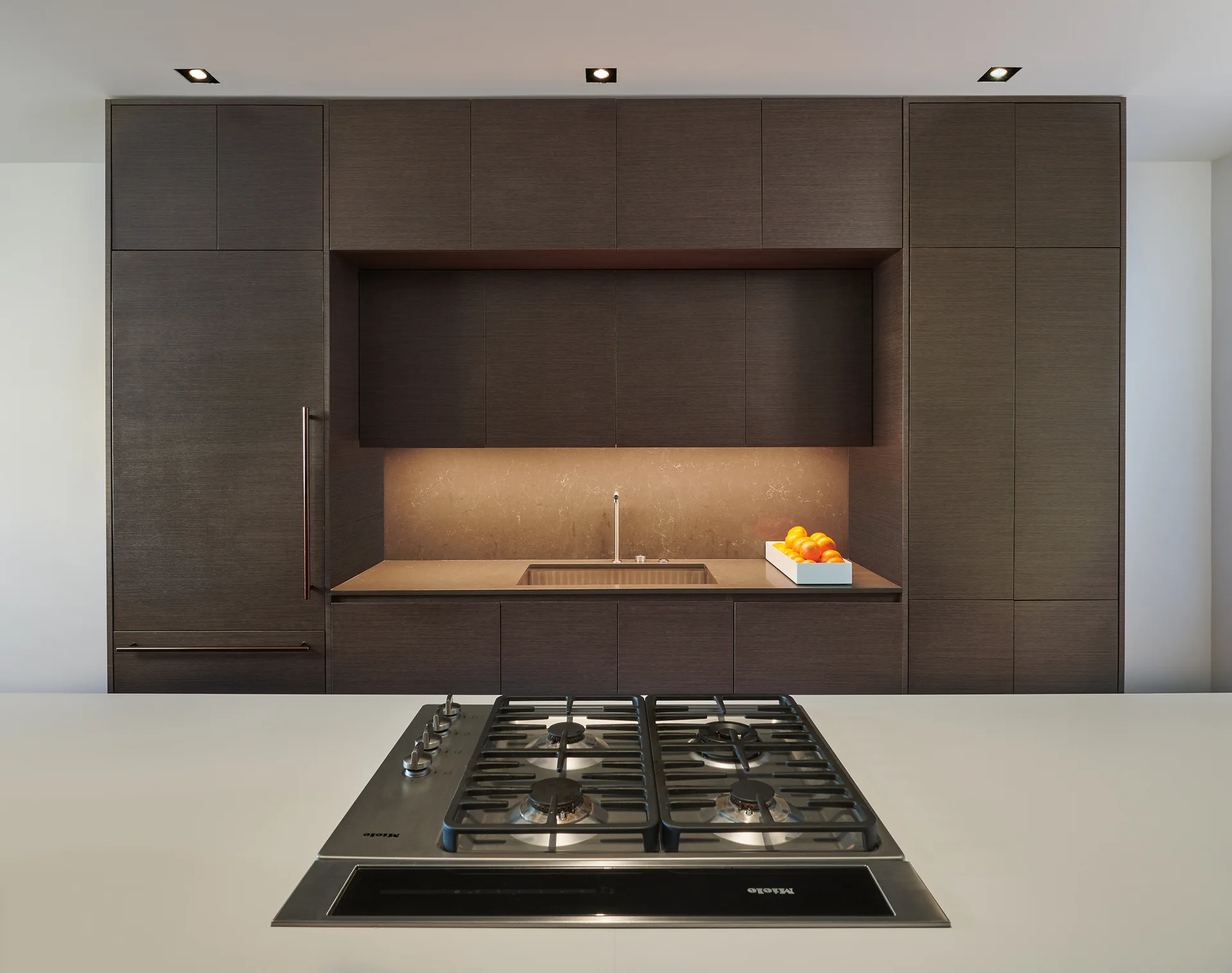 A modern kitchen featuring flat-panel dark brown wood cabinetry, a recessed sink alcove with under-cabinet lighting, and a gas cooktop on a white island.
