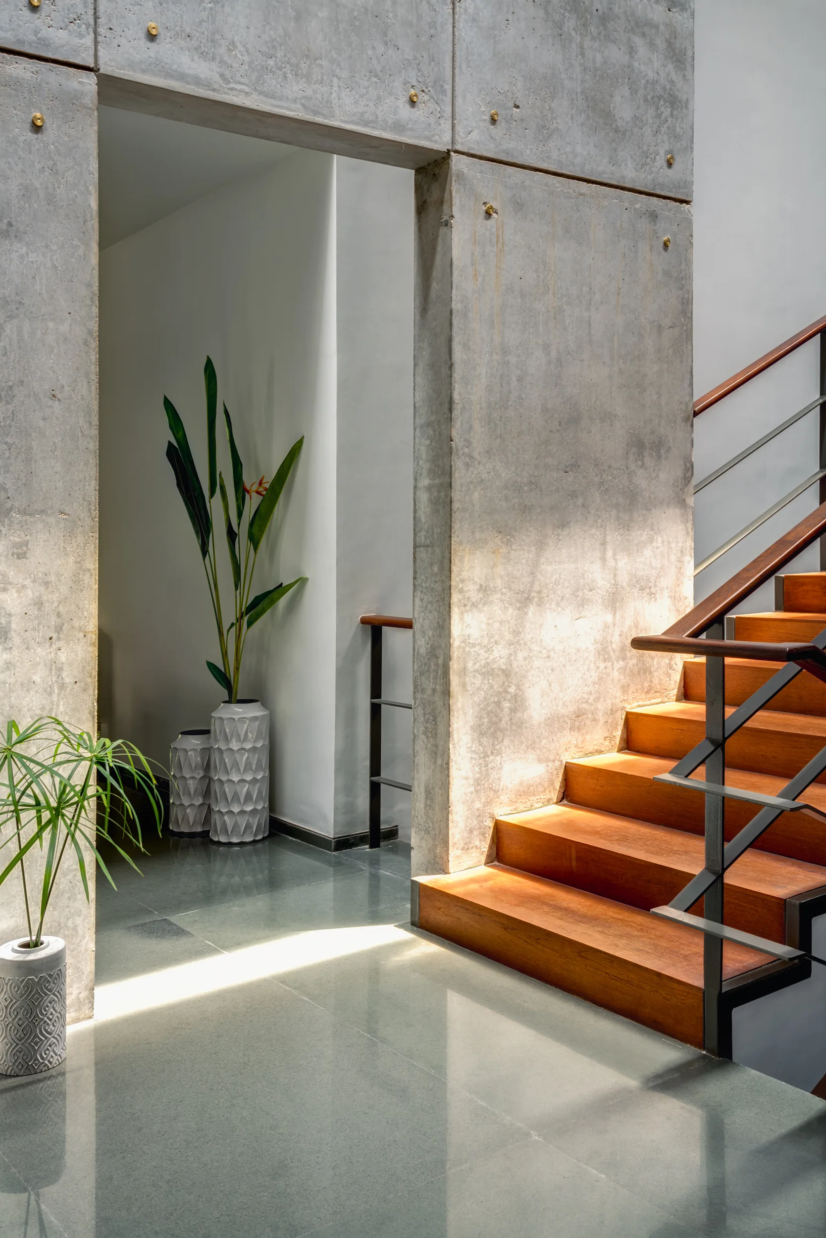 A modern staircase with warm wood treads and a dark metal railing stands next to an architectural concrete wall and polished stone flooring.