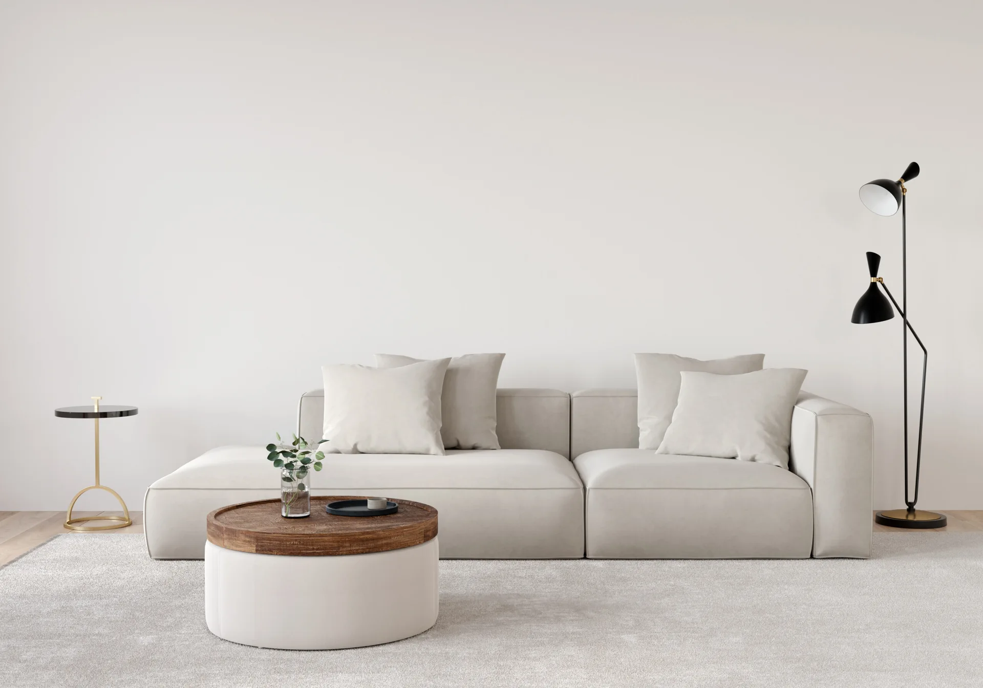A low-profile cream modular sofa sits on a textured grey rug next to a round wooden coffee table and a black metal floor lamp against a blank wall.