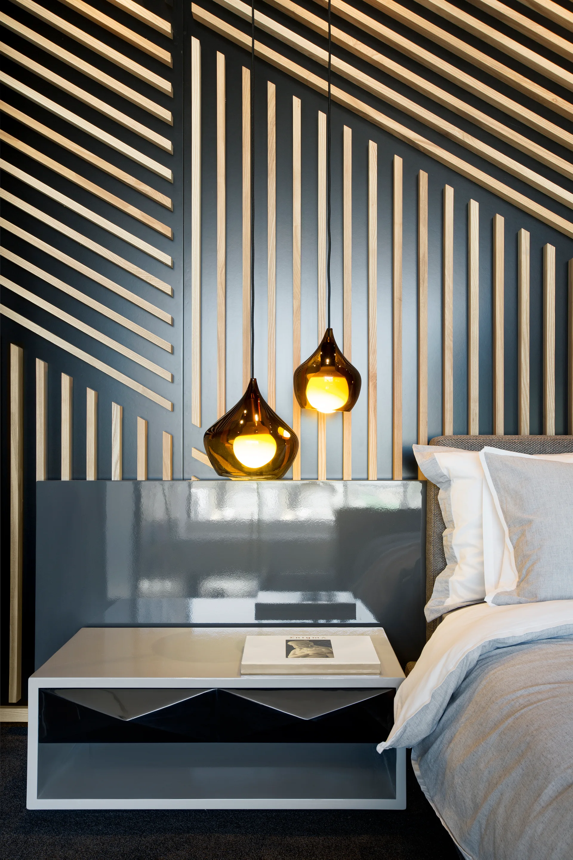 A modern bedroom features a geometric slatted wood accent wall, two low-hanging amber glass pendant lights, and a grey bedside table.
