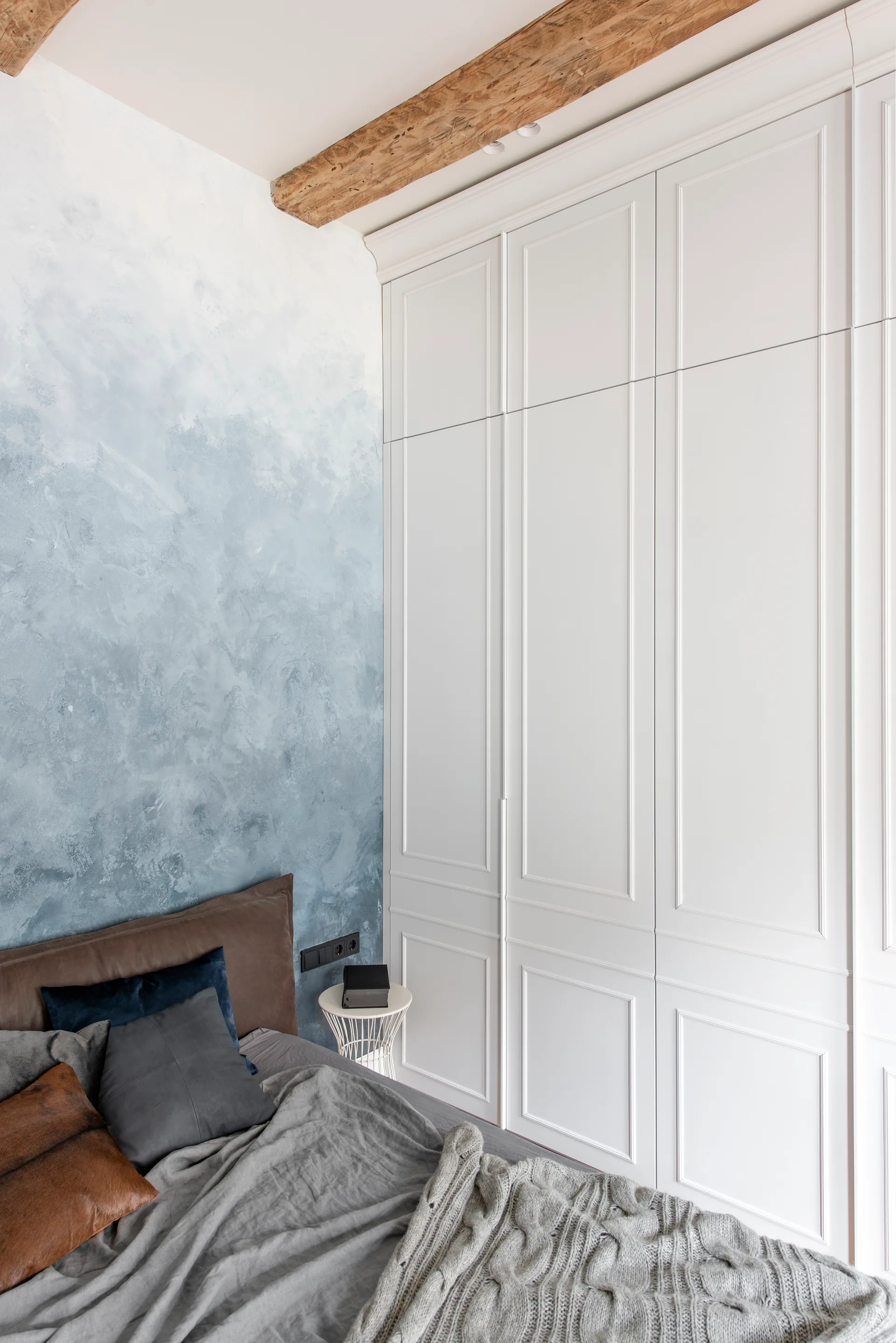 A bedroom features a tall white paneled wardrobe next to a bed with a blue and white ombre textured wall and an exposed wooden ceiling beam.