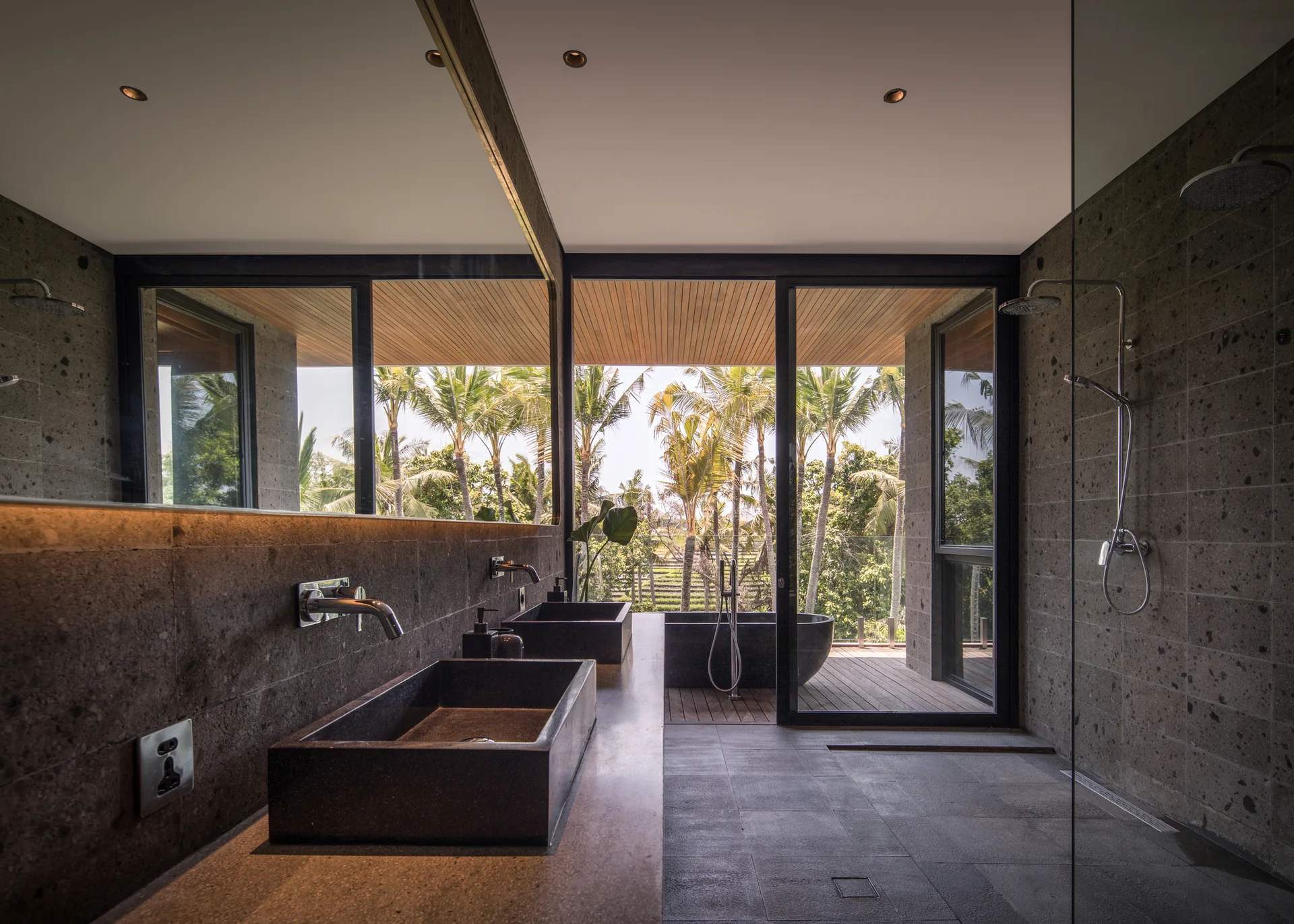 A dark charcoal stone bathroom with a double vanity, sliding glass doors, and a freestanding black bathtub on an outdoor wooden deck.