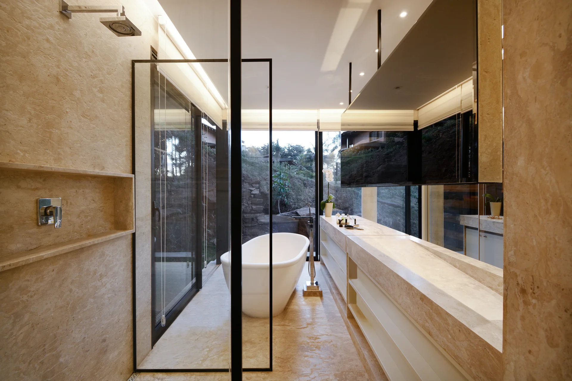 A modern beige stone bathroom features a white freestanding bathtub, a long stone vanity counter, and a floor-to-ceiling glass window overlooking nature.