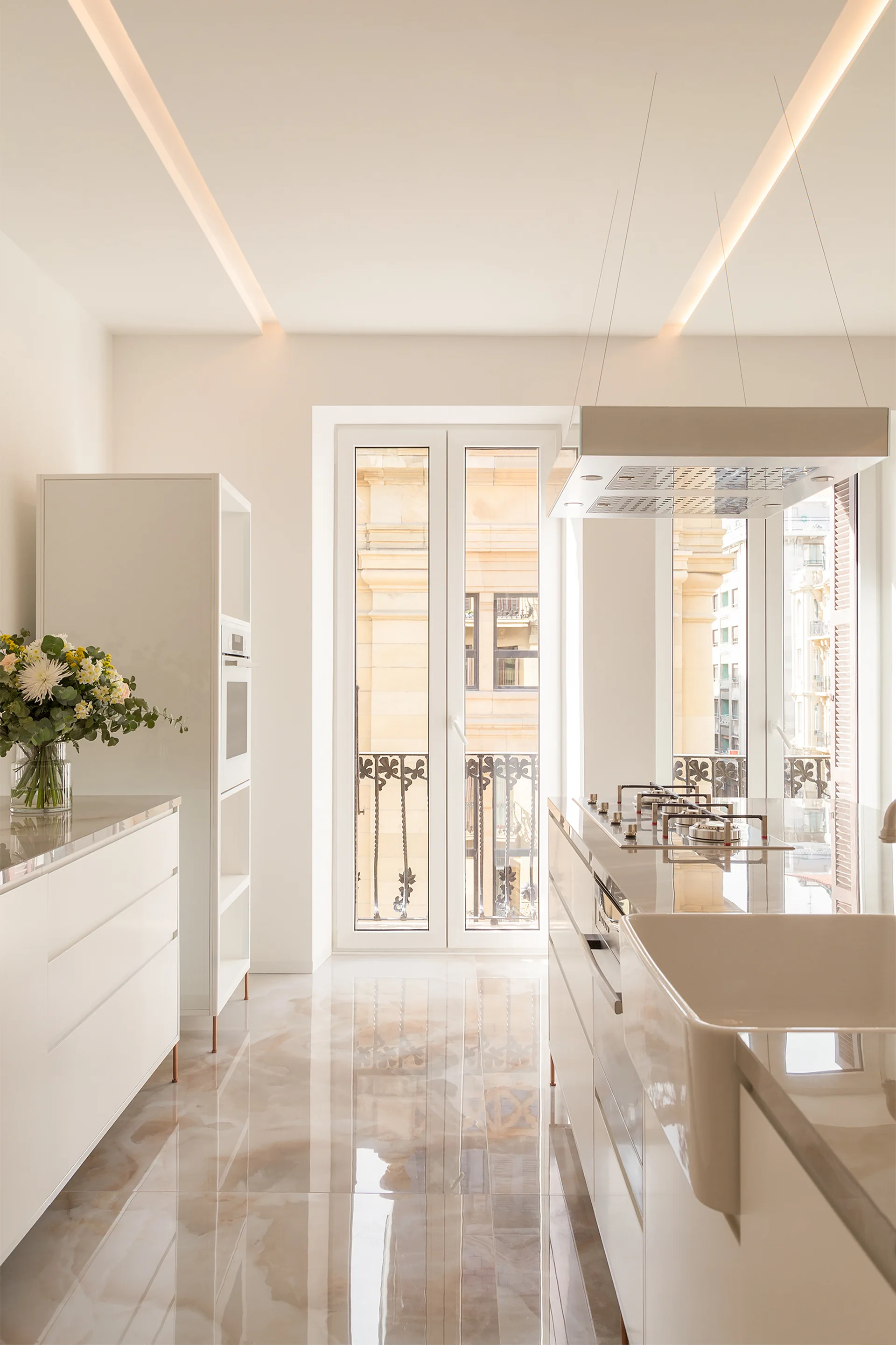 A sleek white kitchen features a central island with an apron-front sink, glossy veined flooring, and tall windows opening to a balcony.